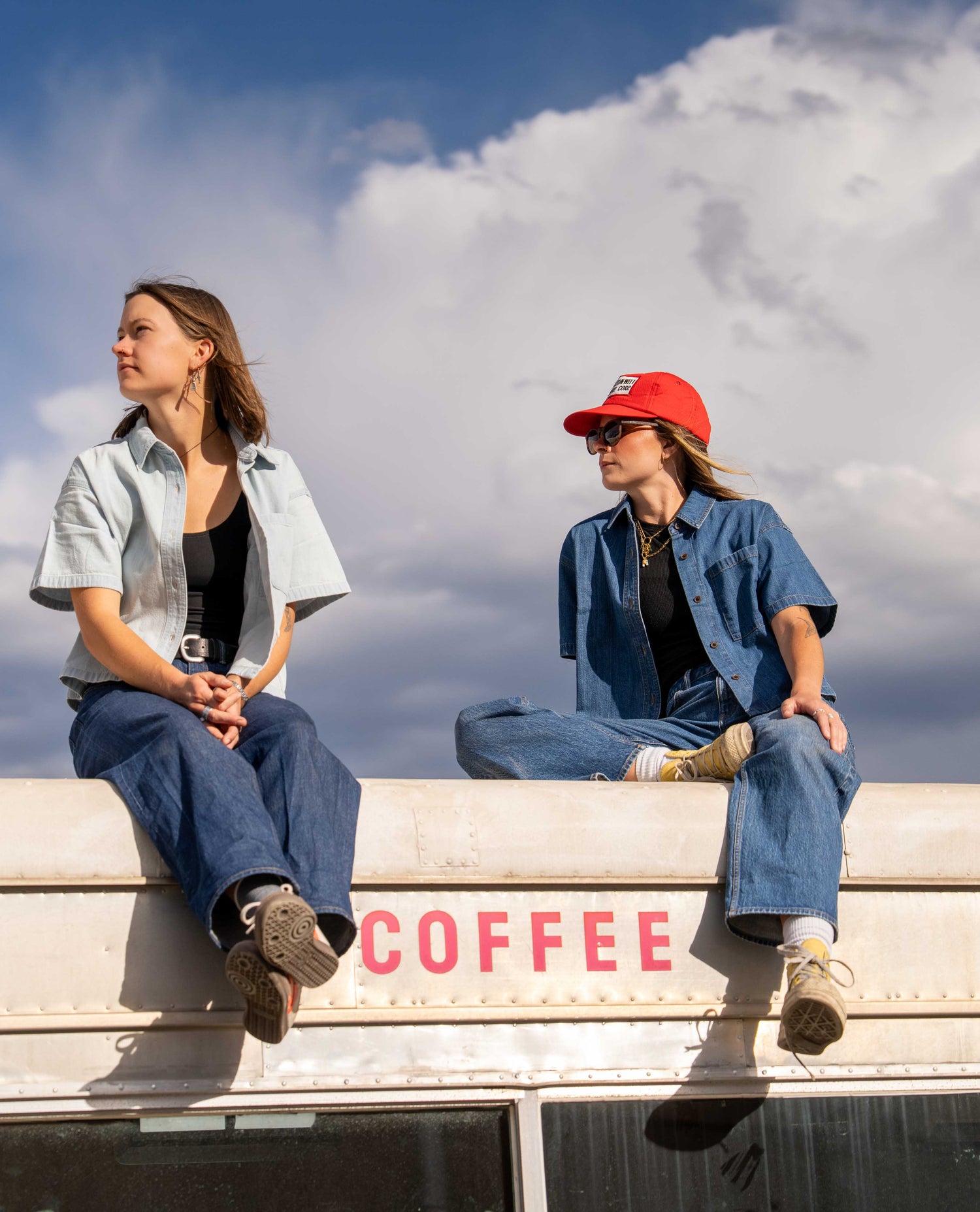 Two women sit on the edge of a vintage white bus under a cloudy blue sky. The woman on the left wears a light blue button-up shirt over a black top and blue jeans.