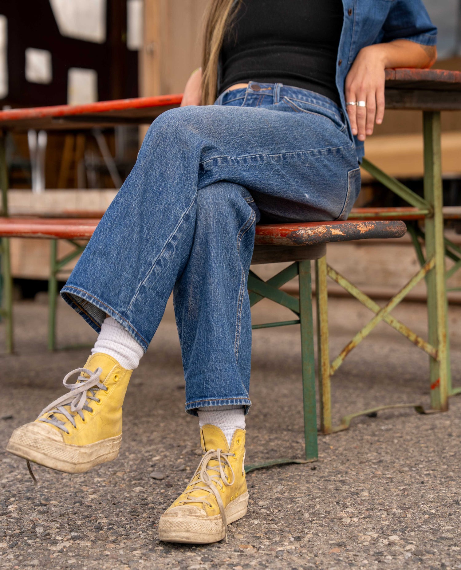 A person sitting on a rustic outdoor bench wearing blue wide-leg jeans, a black top, a blue denim jacket, white socks, and yellow high-top sneakers.