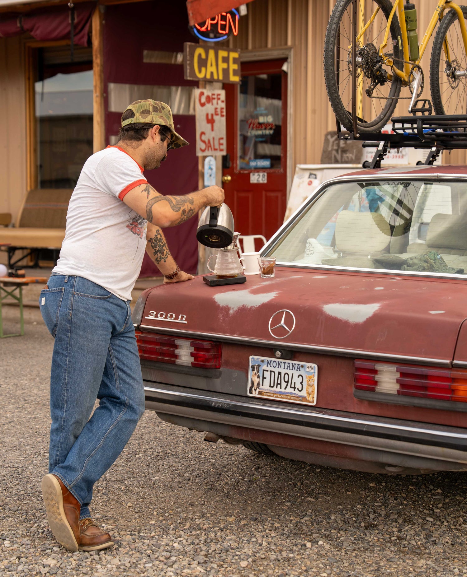 A man in a white graphic t-shirt and blue jeans stands next to a vintage red Mercedes-Benz car, pouring coffee from a black electric coffee maker into a glass mug. The scene is set outdoors in front of a rustic cafe with a bicycle mounted on the car roof rack.