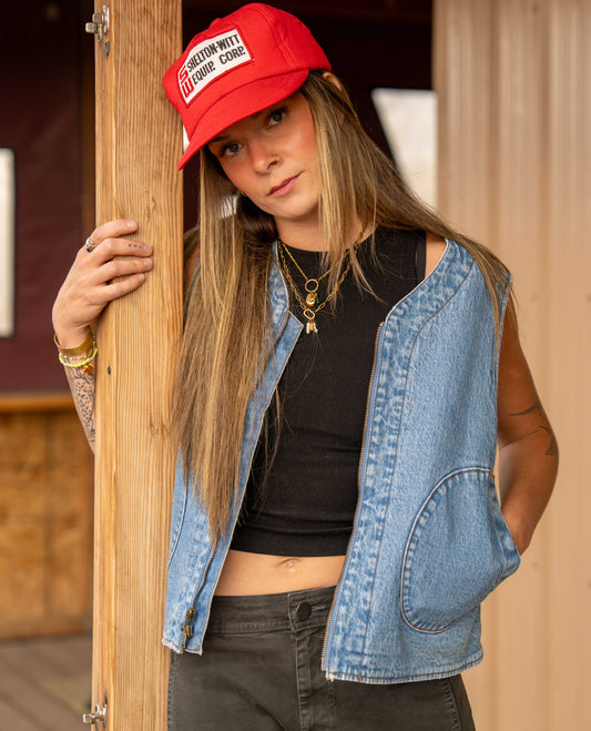 A woman wearing a red baseball cap with a white patch, a light blue denim vest over a black crop top, and dark gray jeans, leaning against a wooden post.