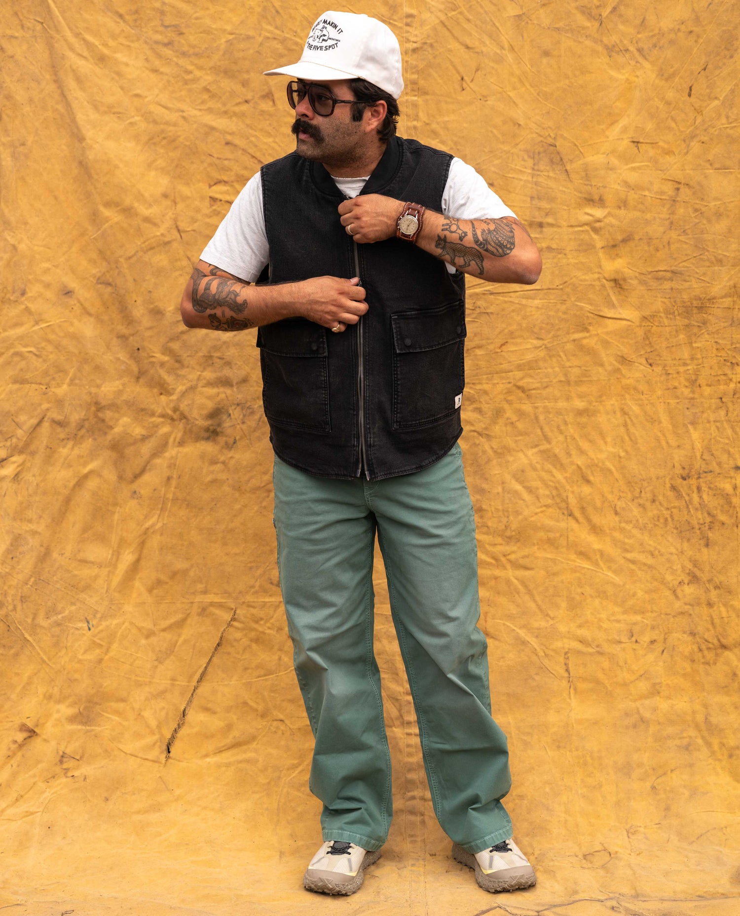 A man wearing a black zippered utility vest over a white t-shirt, olive green pants, and a white cap, standing against a wrinkled yellow backdrop.