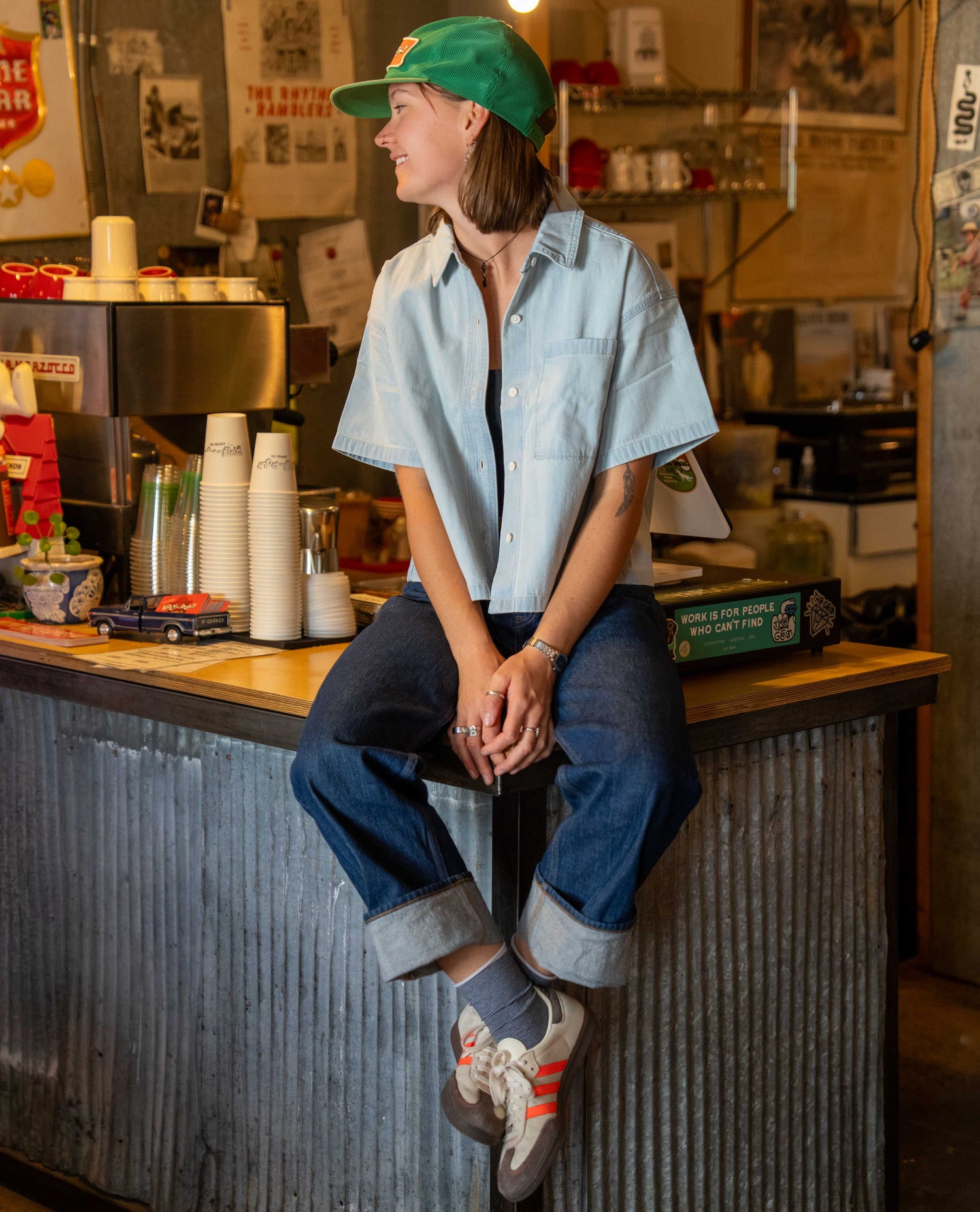 A person sitting on a rustic cafe counter wearing a light blue short-sleeve button-up shirt, dark blue jeans, a green baseball cap, and white sneakers with orange stripes, in a warm, vintage-style coffee shop interior.
