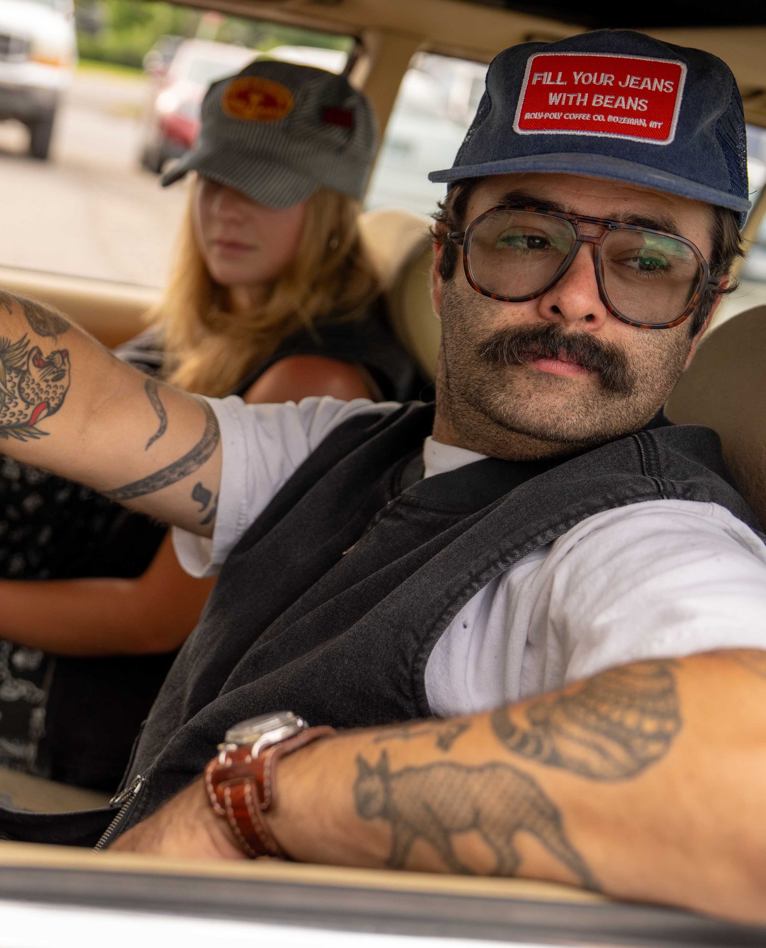 A man in a car wearing a dark gray vest, white t-shirt, glasses, and a blue cap with a red and white patch that reads 'FILL YOUR JEANS WITH BEANS'. A woman is seated in the background.
