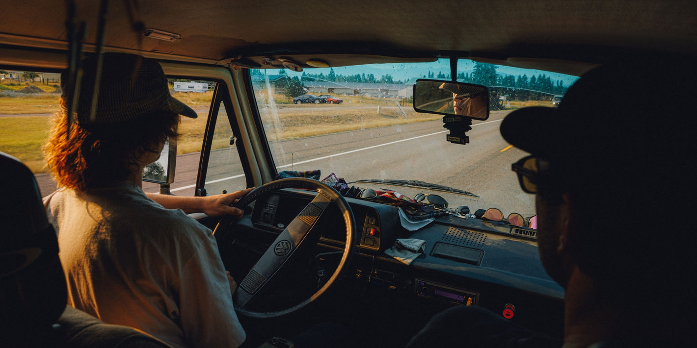 A person driving a vehicle on a road, seen from inside the cabin with another person in the foreground. The scene is backlit by warm sunlight, and the windshield shows an outdoor landscape.