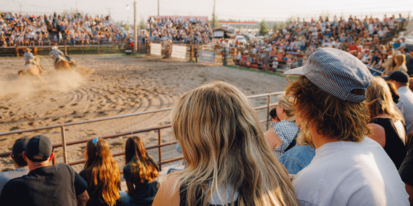 A crowd of spectators watches a rodeo event in an outdoor arena, with a rider on horseback kicking up dust. The foreground shows the backs of several audience members, including one in a blue-and-white striped cap and another with long blonde hair.