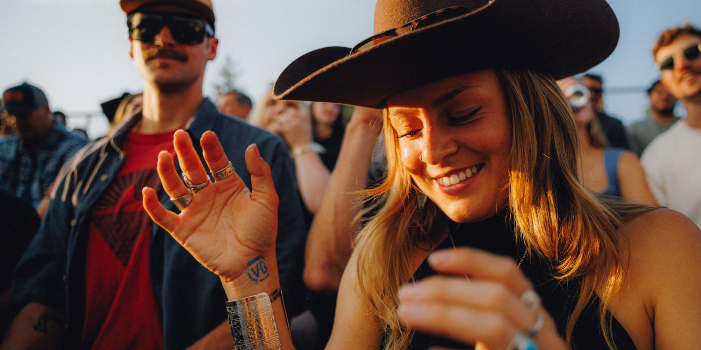 A smiling woman in a wide-brimmed brown cowboy hat waves to a crowd at an outdoor festival during golden hour, with other attendees blurred in the background.