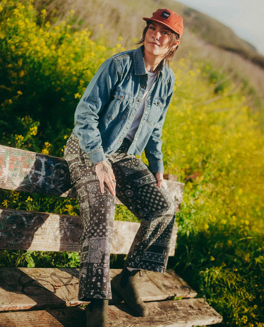 A person sitting on a weathered wooden bench outdoors in a grassy field with yellow flowers. They are wearing a blue denim shirt, a red hat, dark patterned pants, and dark boots.