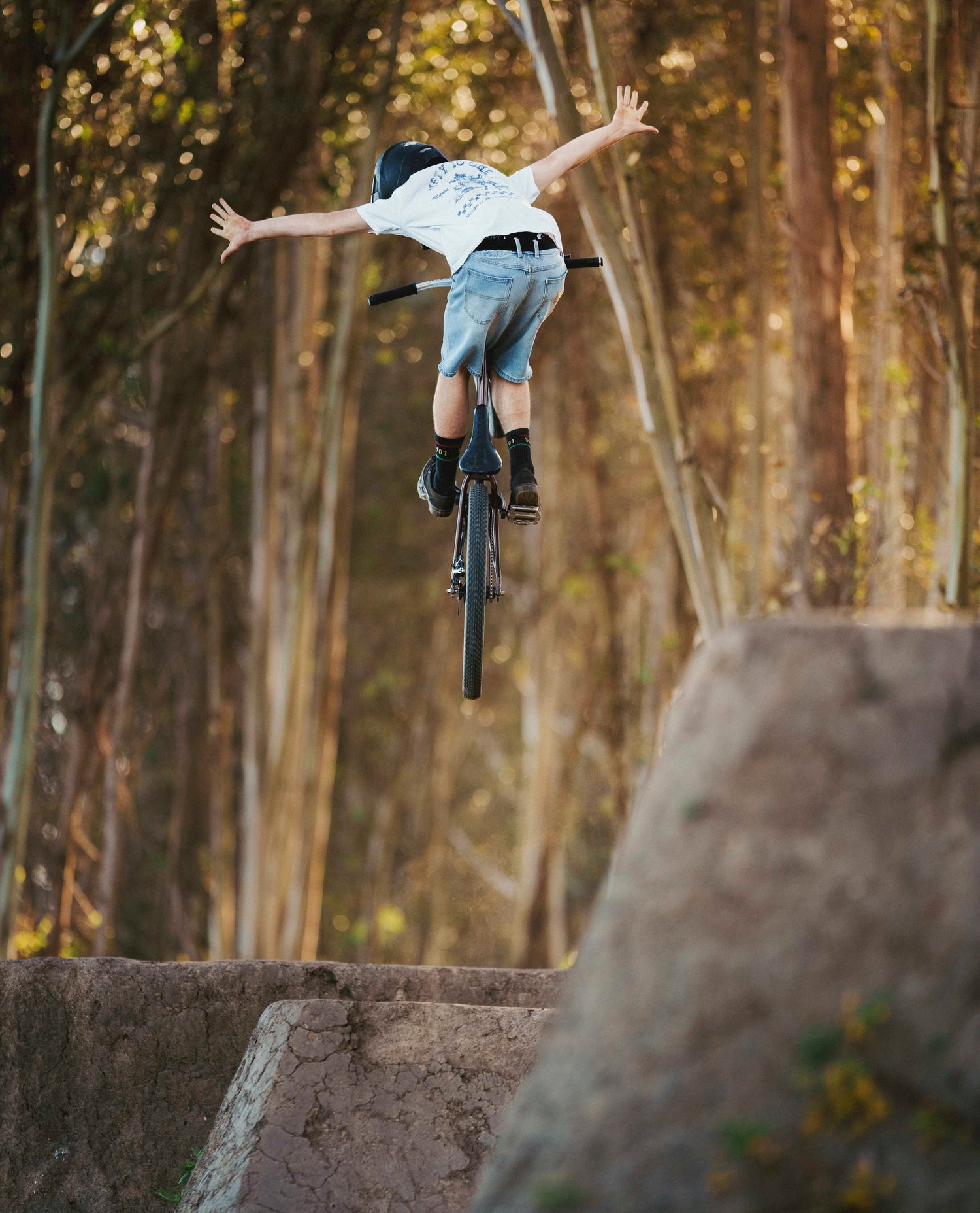 Young man in a white t shirt and blue jean shorts with black high socks jumping a bicycle in the woods high in the air. 