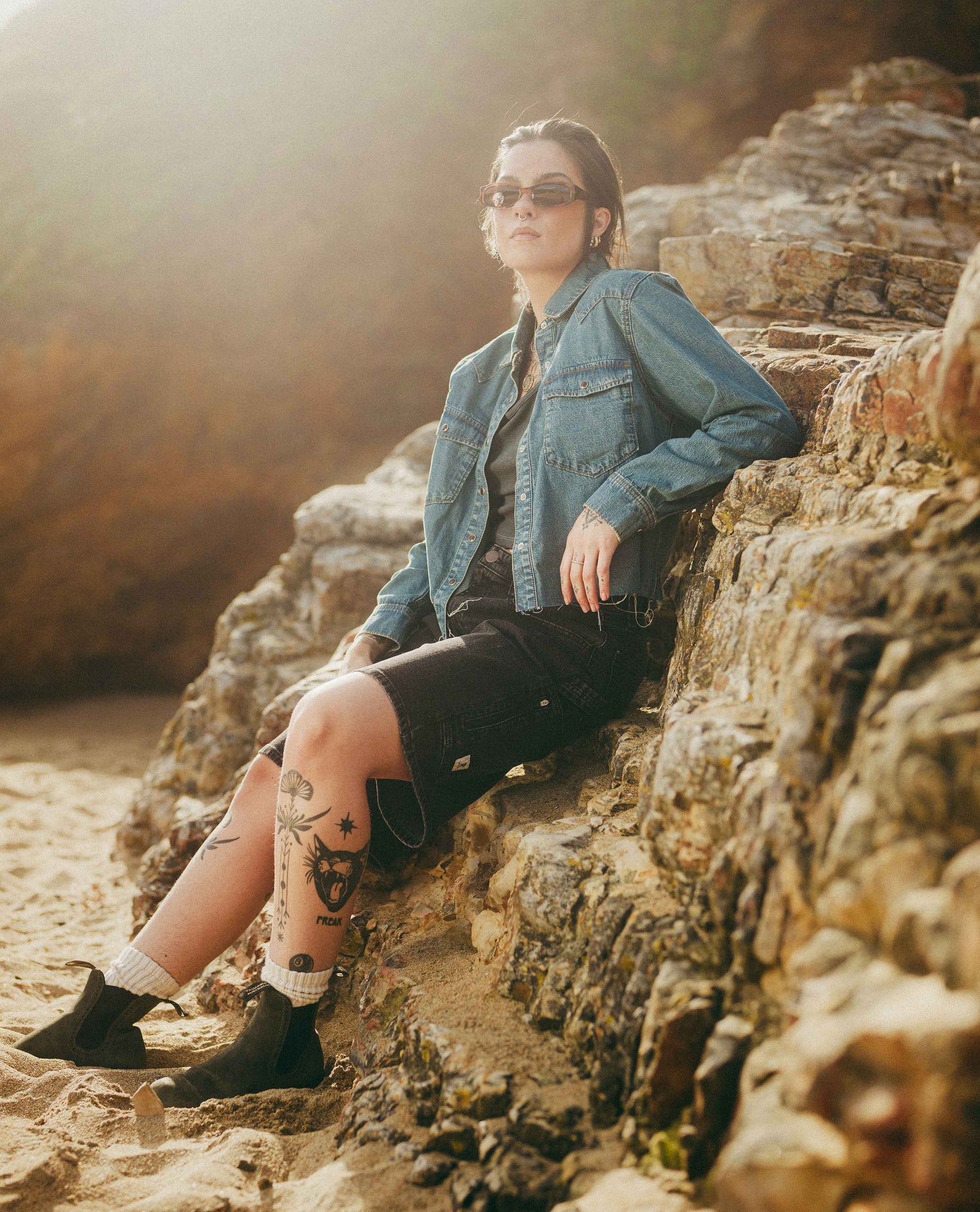 A woman wearing a blue denim shirt, black shorts, and black boots sits on a rocky beach with warm sunlight filtering through the background.