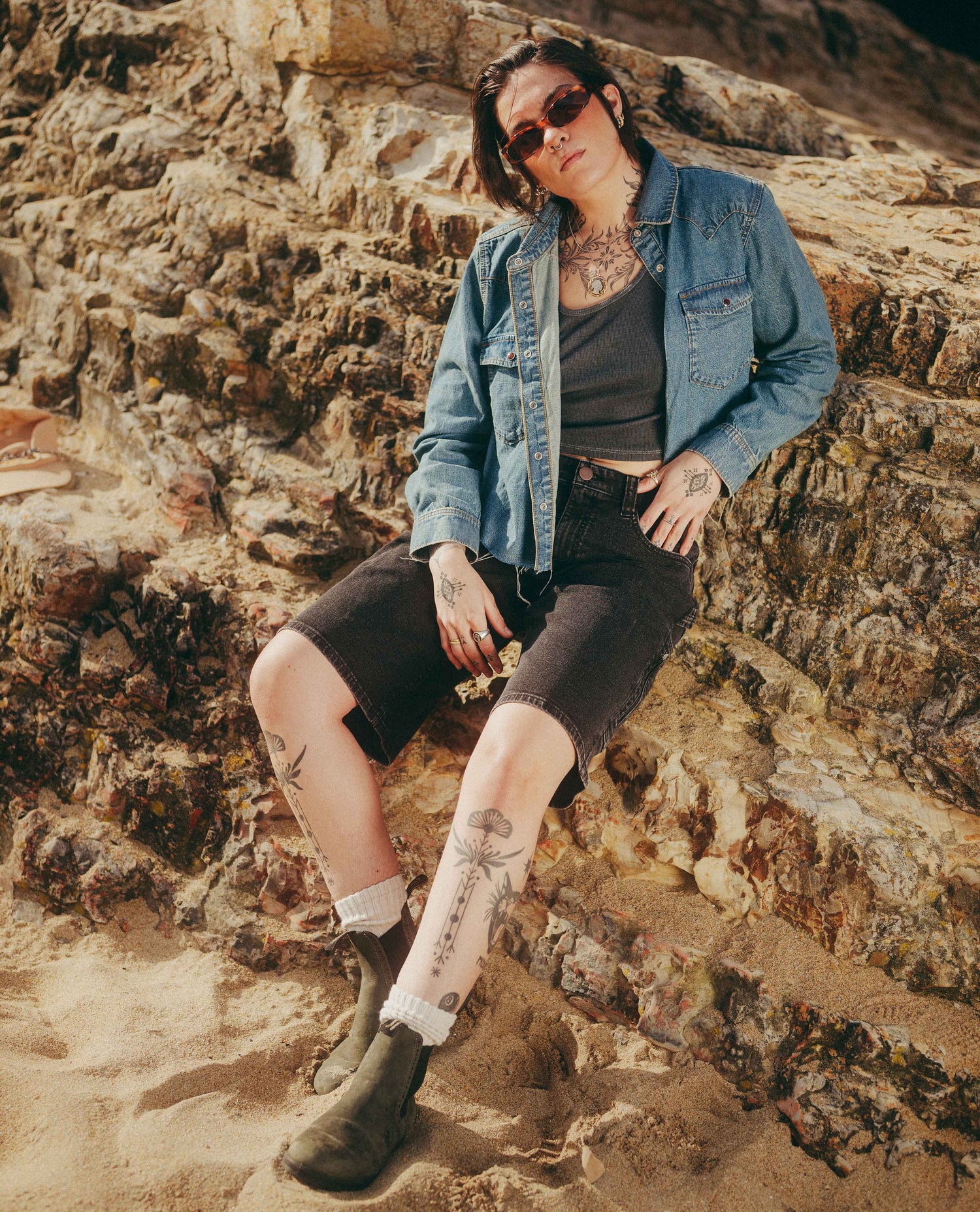 A person sits on a sandy rock formation wearing a light blue denim shirt, a dark gray tank top, black denim shorts, white socks, and dark boots.