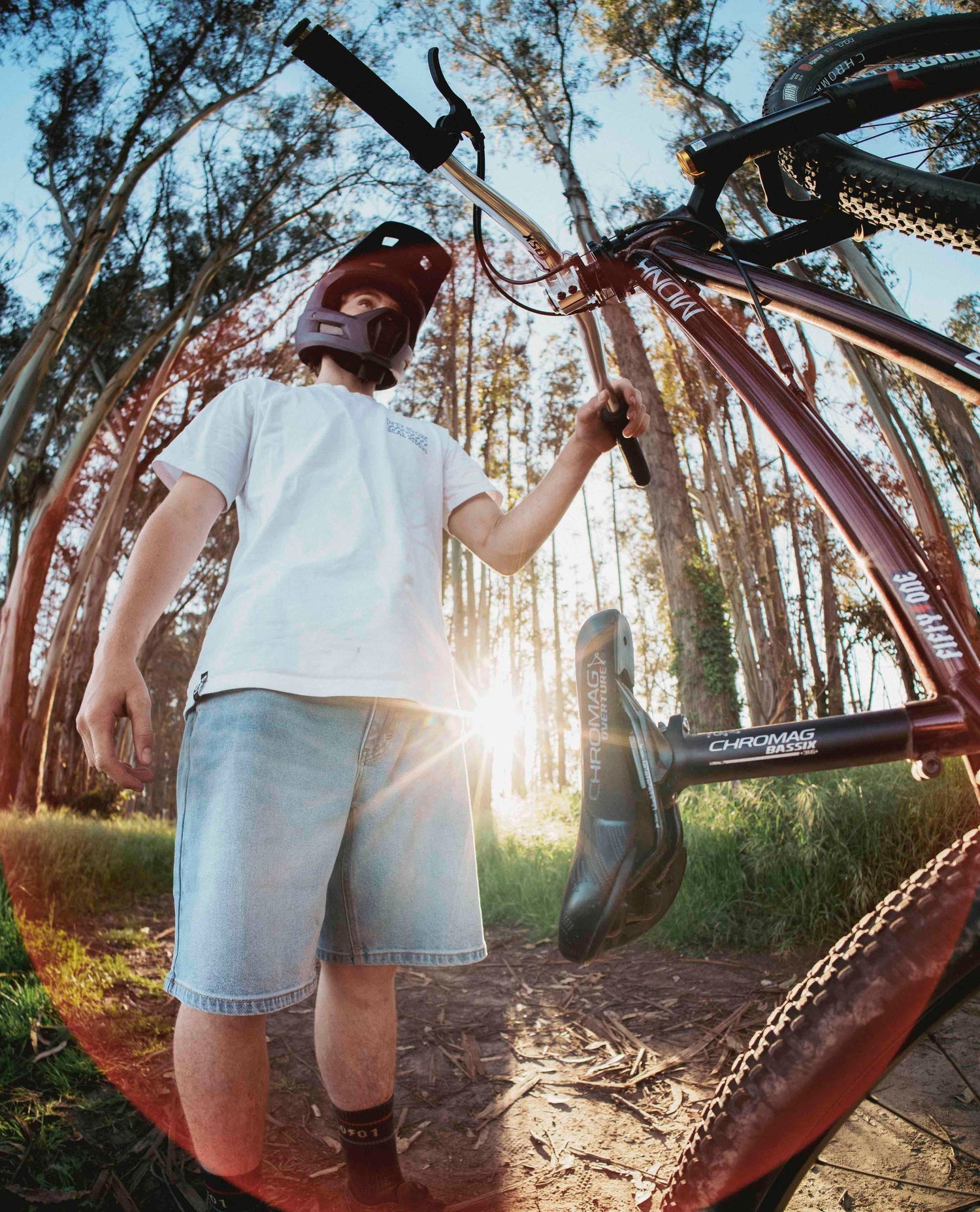 A wide angle photo of a young man holding a white t shirt and a pair of light blue jean shorts. He is holding a bike in his left hand. 