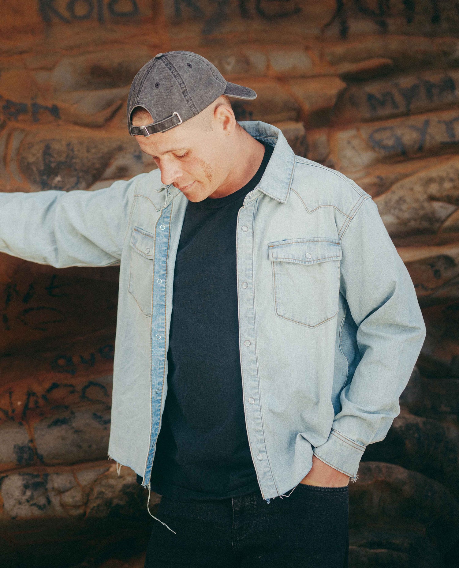 A man wearing a light-wash denim button-up shirt over a black t-shirt and a backwards dark gray baseball cap, standing against a backdrop with graffiti.