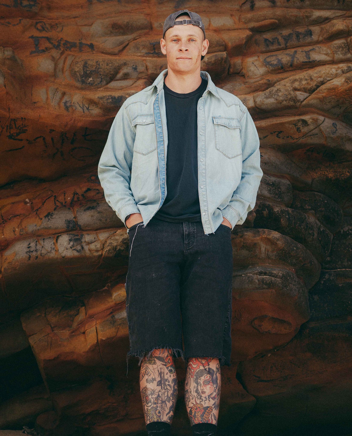A man stands in front of a rocky, graffiti-covered backdrop wearing a light blue denim shirt over a black t-shirt, black denim shorts, and a backwards dark baseball cap.
