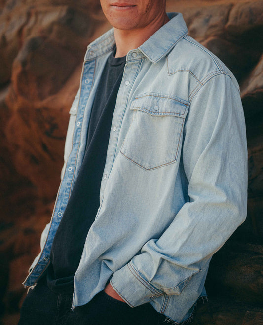 A man wearing a light blue denim button-up shirt over a black t-shirt, posed against a warm, rocky background.