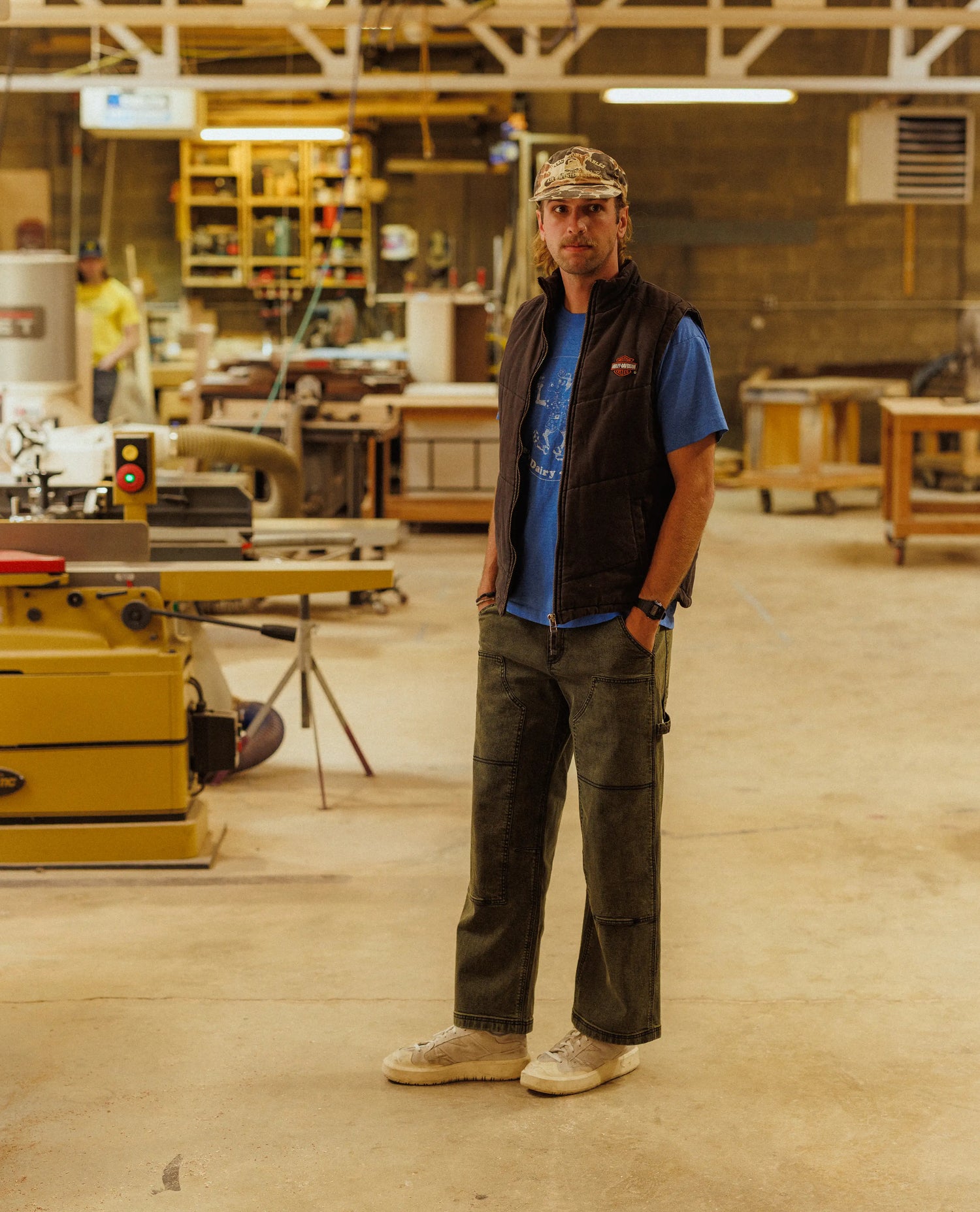 Man standing in a workshop wearing a blue shirt, black vest, and green pants.