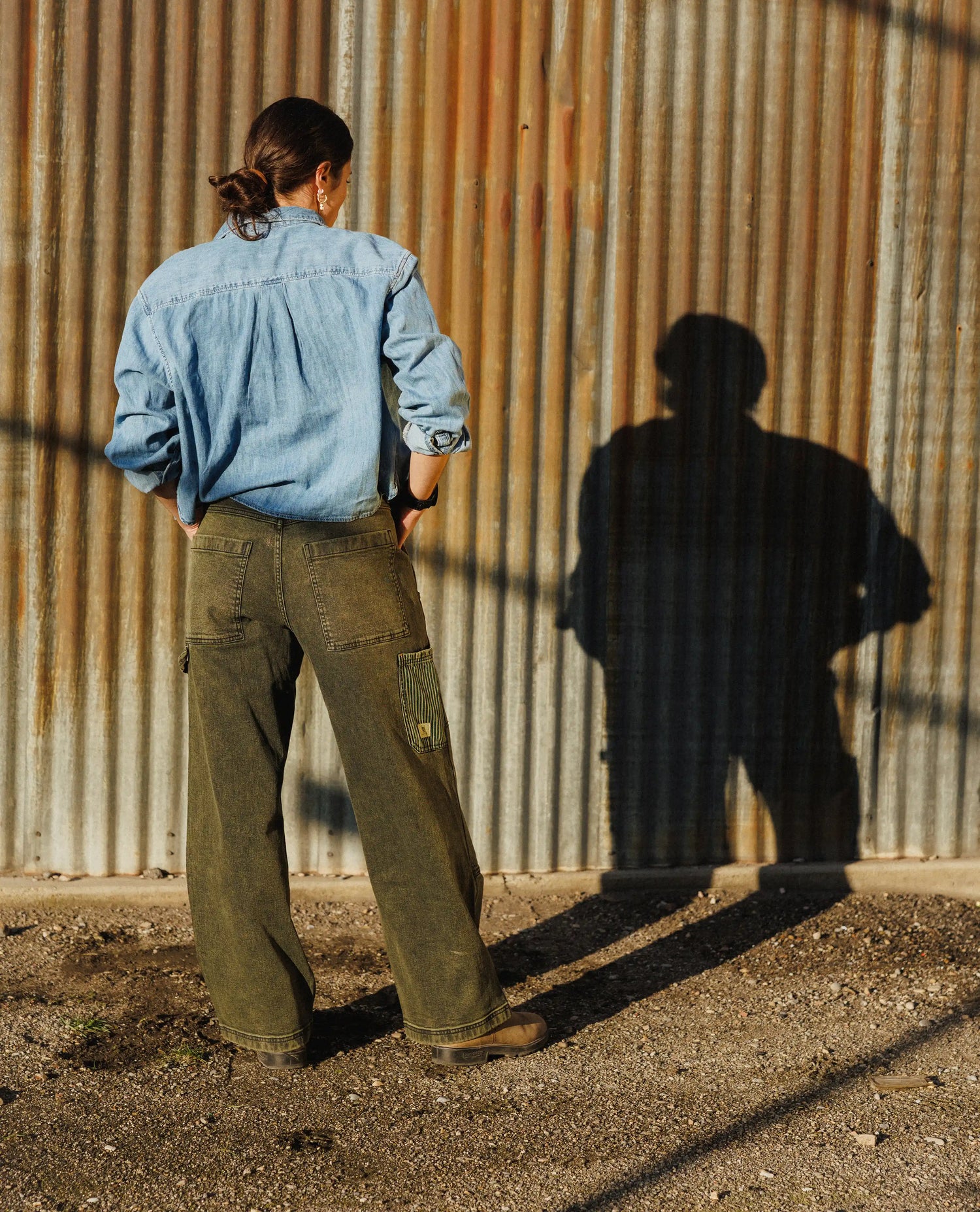 Person wearing a denim shirt and green pants standing in front of a corrugated metal wall.