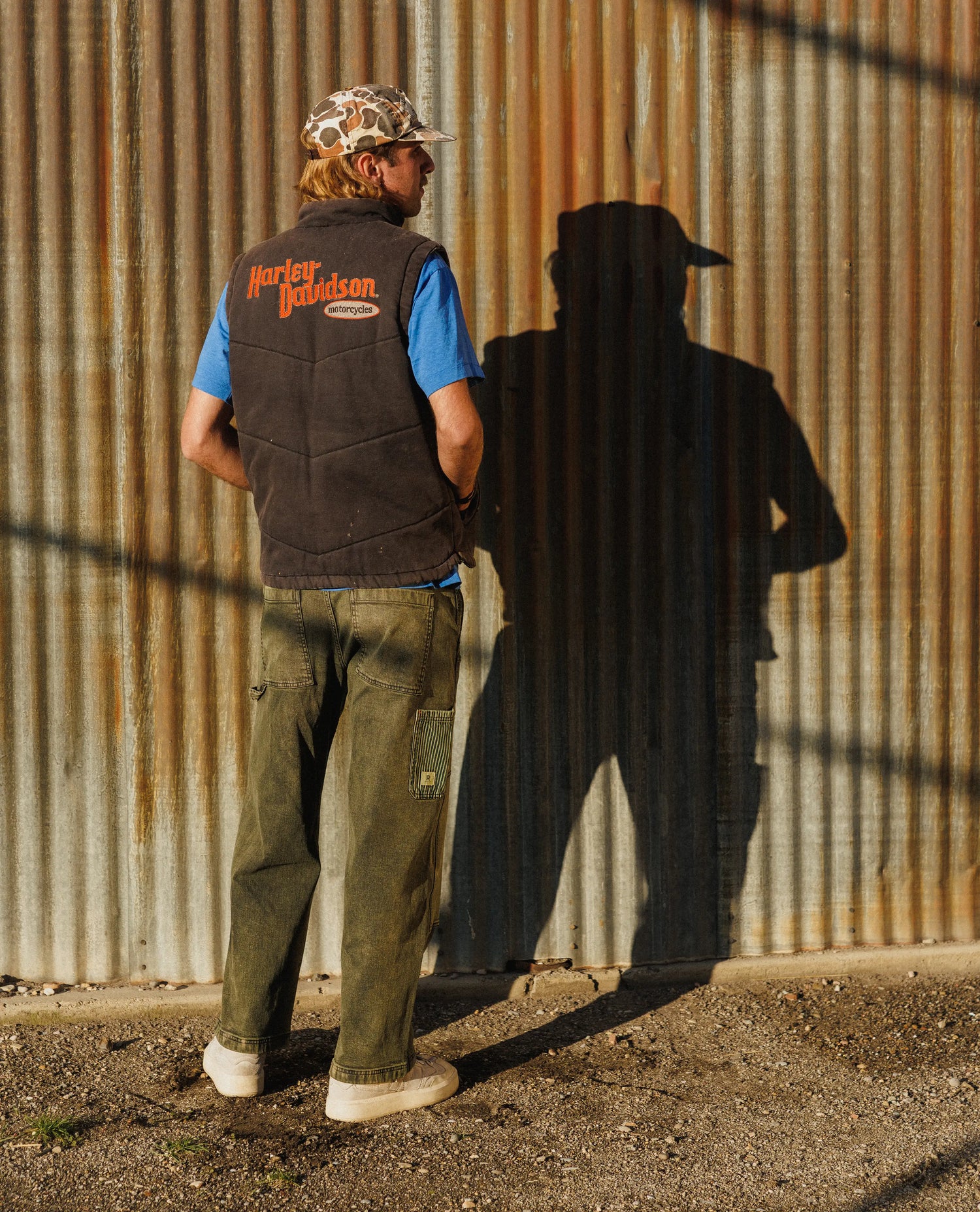 Man wearing a Harley Davidson vest standing against a rusty corrugated metal wall.