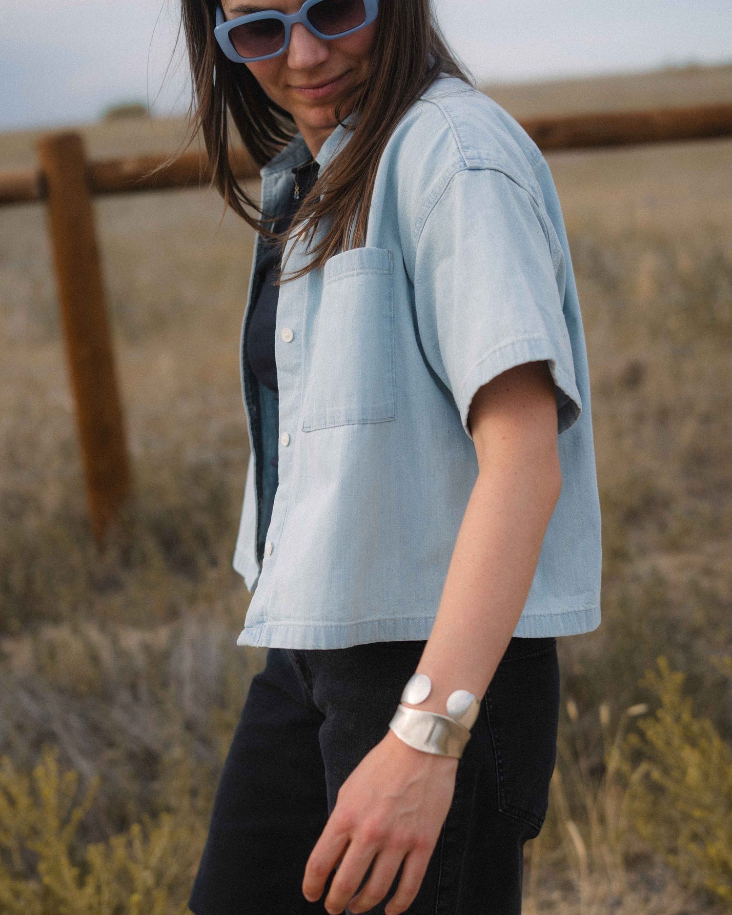 A woman wearing a light blue cropped short-sleeve button-up shirt over a dark top and black pants, standing outdoors in a dry grassy field.