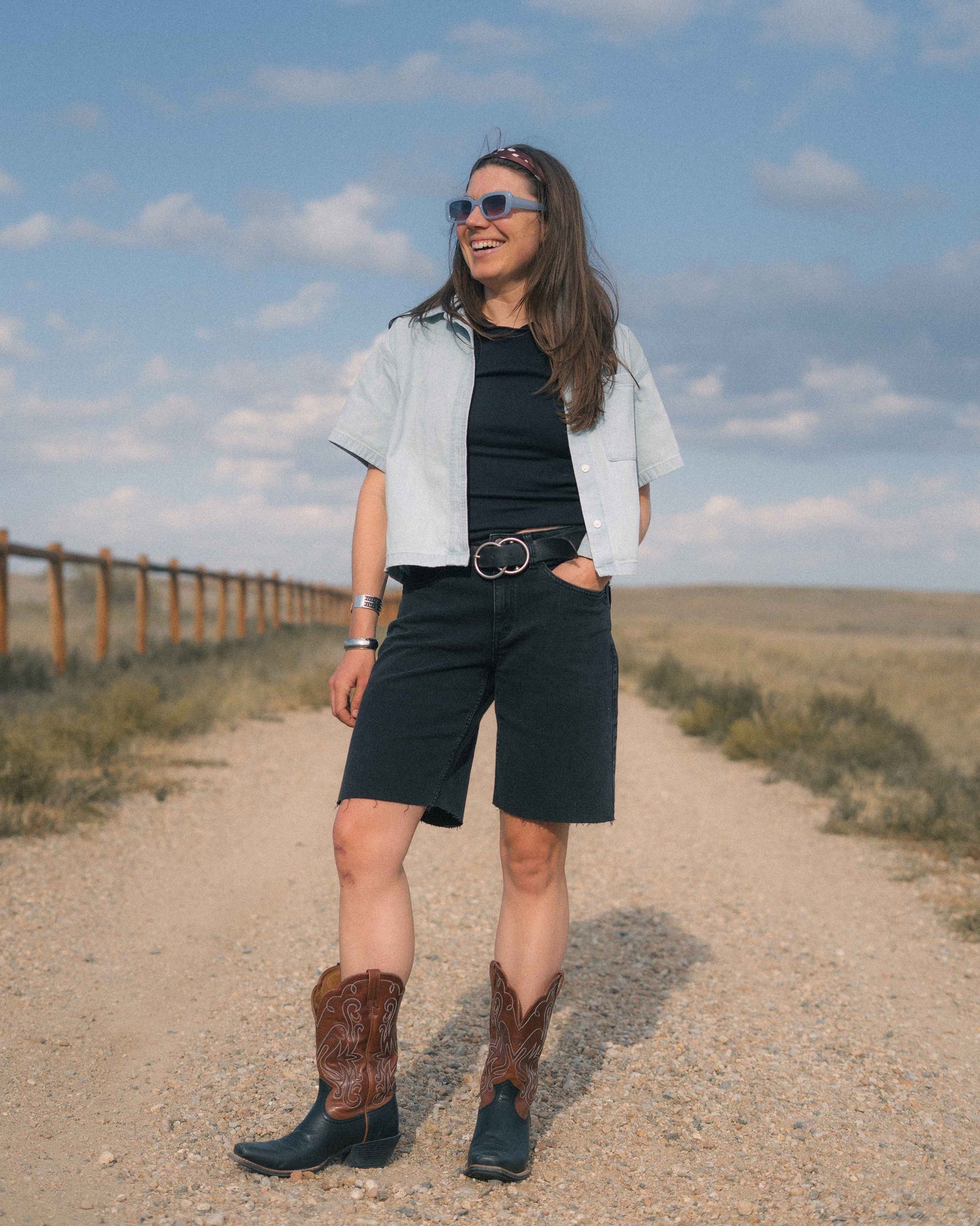 A woman stands on a dirt path outdoors wearing a light blue short-sleeve button-up shirt over a black top, black shorts, a black belt with a silver buckle, and dark cowboy boots with brown shaft detailing. She is wearing sunglasses, and the background shows a grassy field under a blue sky with clouds.