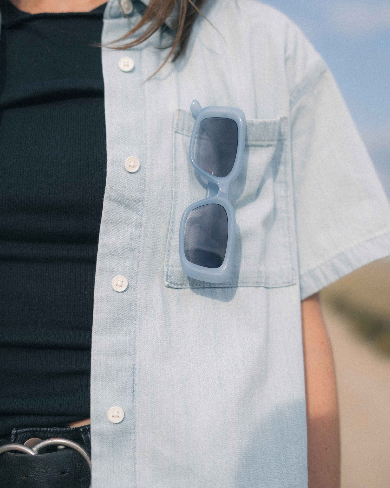 A close-up of a person wearing a light blue short-sleeve button-up shirt over a black ribbed top, with a pair of light blue sunglasses resting in the shirt's chest pocket.