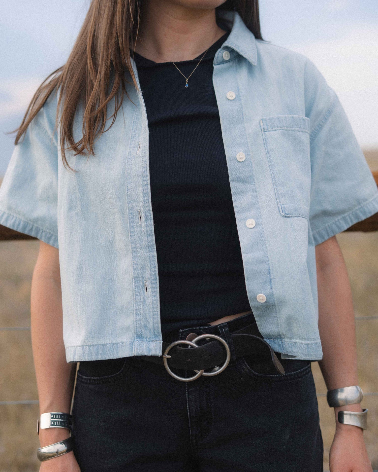A person wearing a light blue cropped short-sleeve button-up shirt over a black top and dark jeans with a double-ring belt, photographed outdoors.