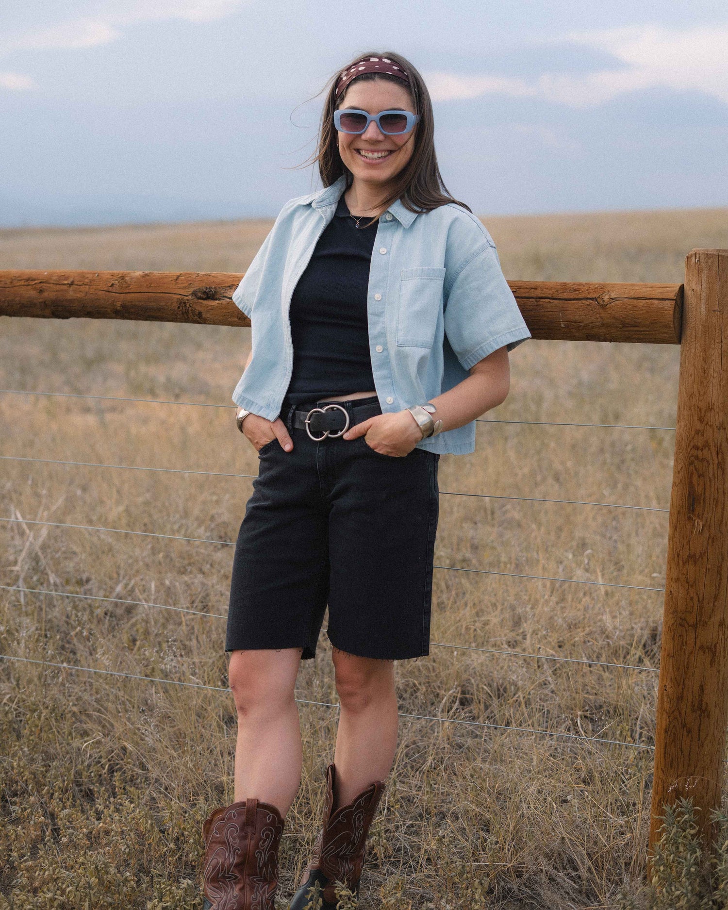 A woman stands outdoors in a grassy field wearing a light blue short-sleeve button-up shirt over a black top, black shorts, a black belt with a silver buckle, brown cowboy boots, and sunglasses.