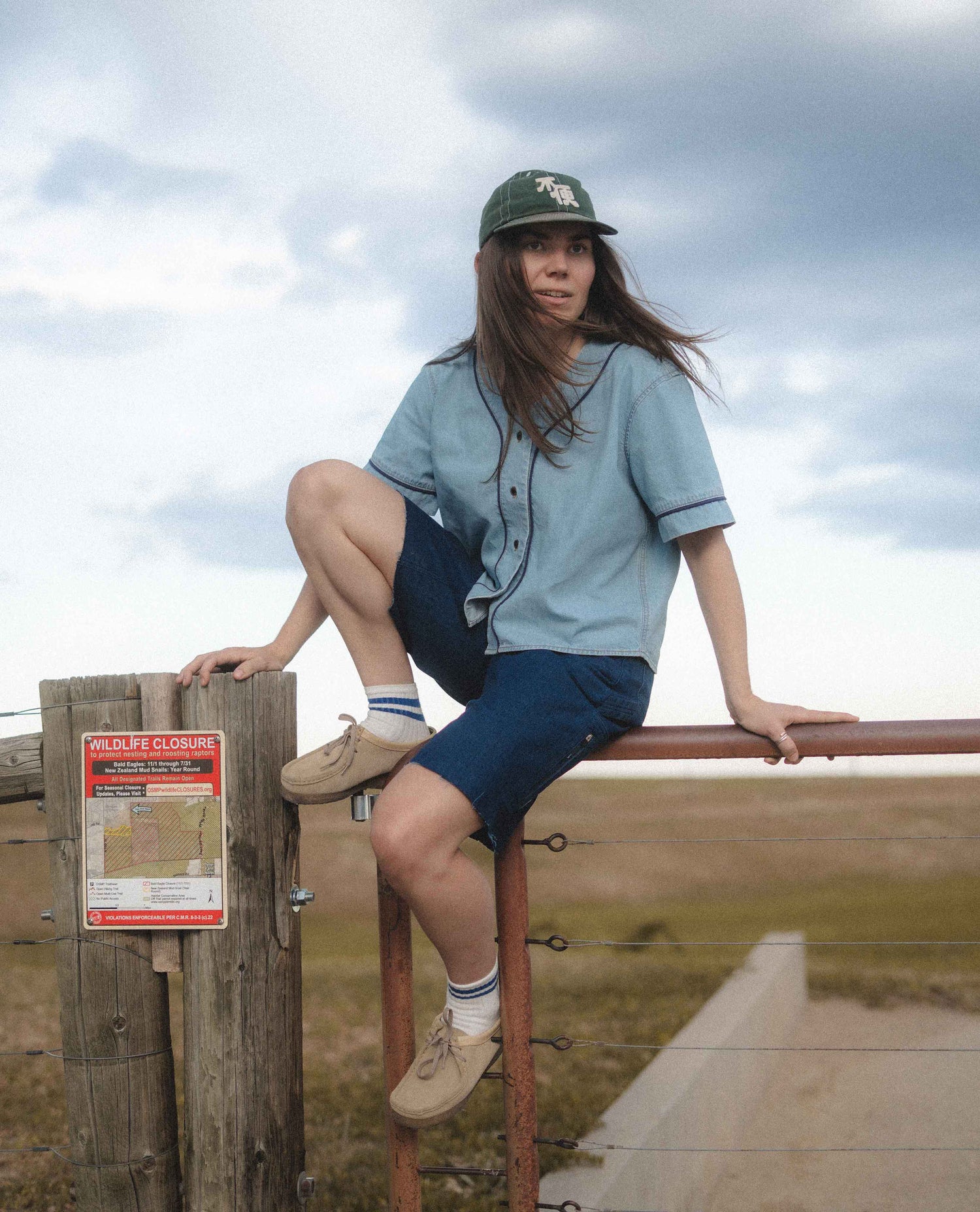 A woman wearing a light blue short-sleeve button-up shirt, navy shorts, beige sneakers, and a dark green baseball cap sits on a wooden fence post outdoors under a cloudy sky.