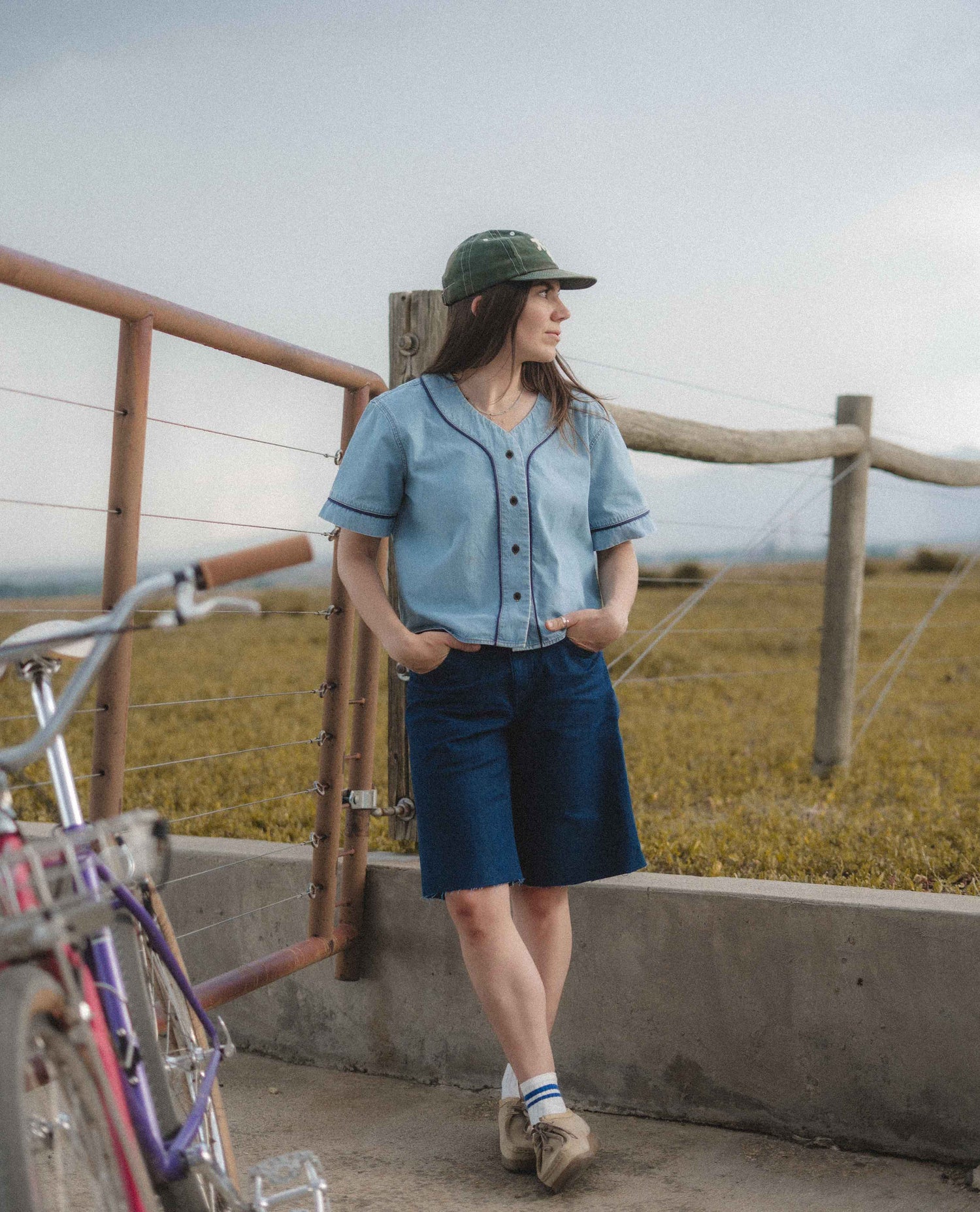 A woman stands outdoors near a fence and field, wearing a light blue short-sleeve button-front shirt, dark blue shorts, a green cap, white socks, and tan shoes. A bicycle is partially visible in the foreground.