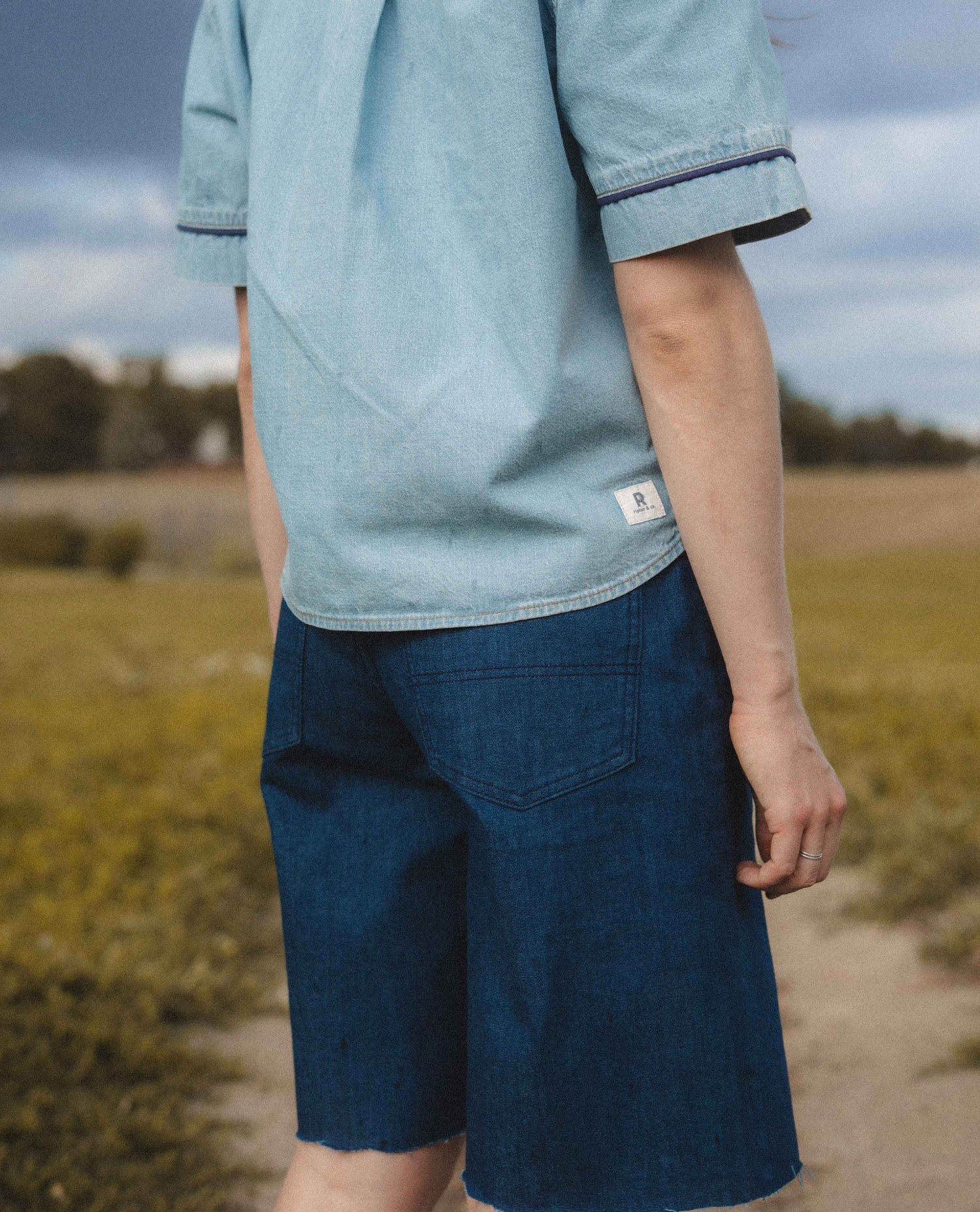 A person outdoors wearing a light blue chambray short-sleeve shirt and dark blue denim shorts, photographed from the back with a blurred field and cloudy sky in the background.