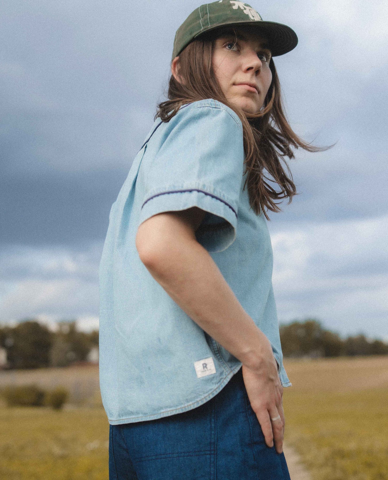 A woman wearing a light blue short-sleeve denim shirt with dark piping on the sleeve and a green baseball cap, standing outdoors in a grassy field under a cloudy sky.