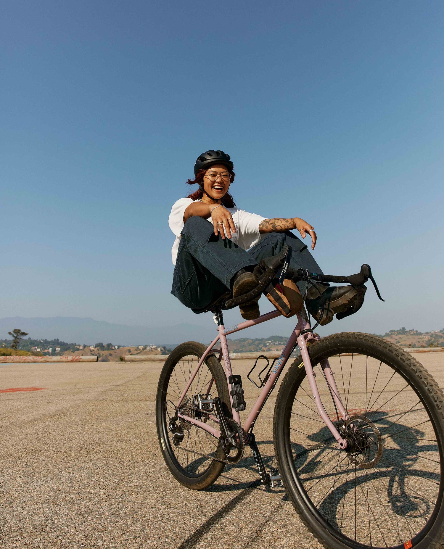 A woman grins as she playfully rides her bike with her legs on the handlebar while sitting on the bike's seat. She is wearing a helmet, a white t shirt, and blue pants. Her bike is pink with large knobby tires. 