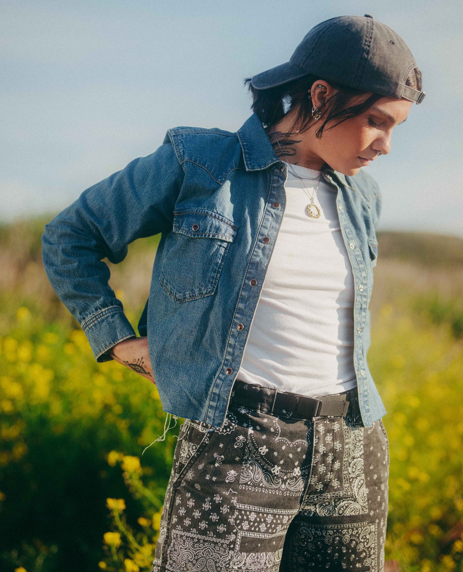 A person standing outdoors in a field of yellow flowers, wearing a blue denim jacket over a white t-shirt, patterned pants, and a dark baseball cap.