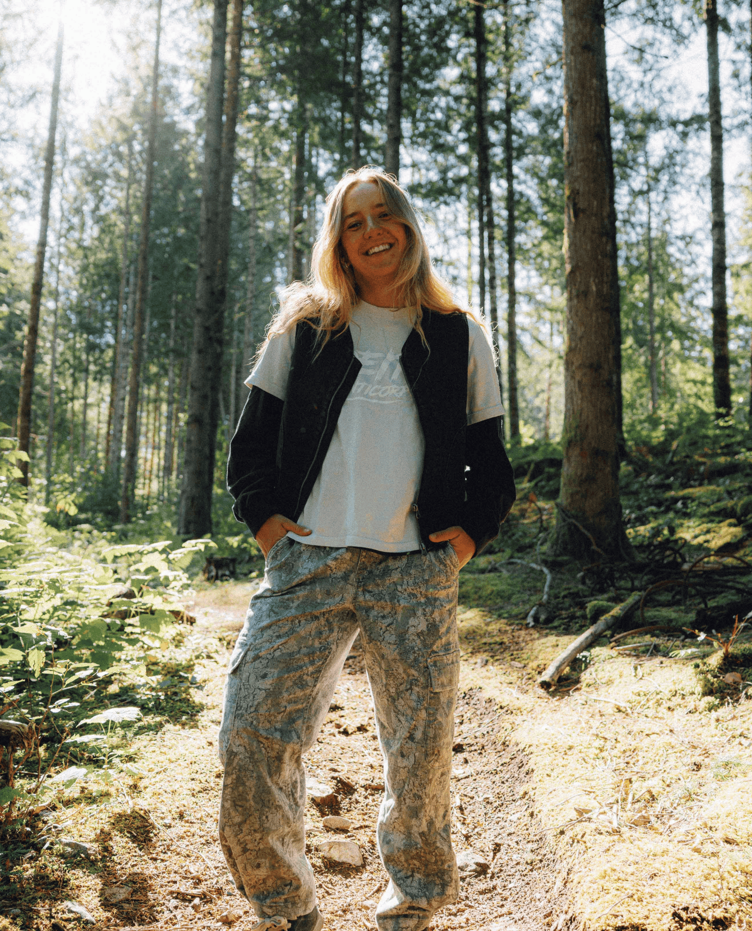 A smiling woman stands on a sunlit forest path wearing a black vest, a gray graphic T-shirt, and loose camouflage jeans.
