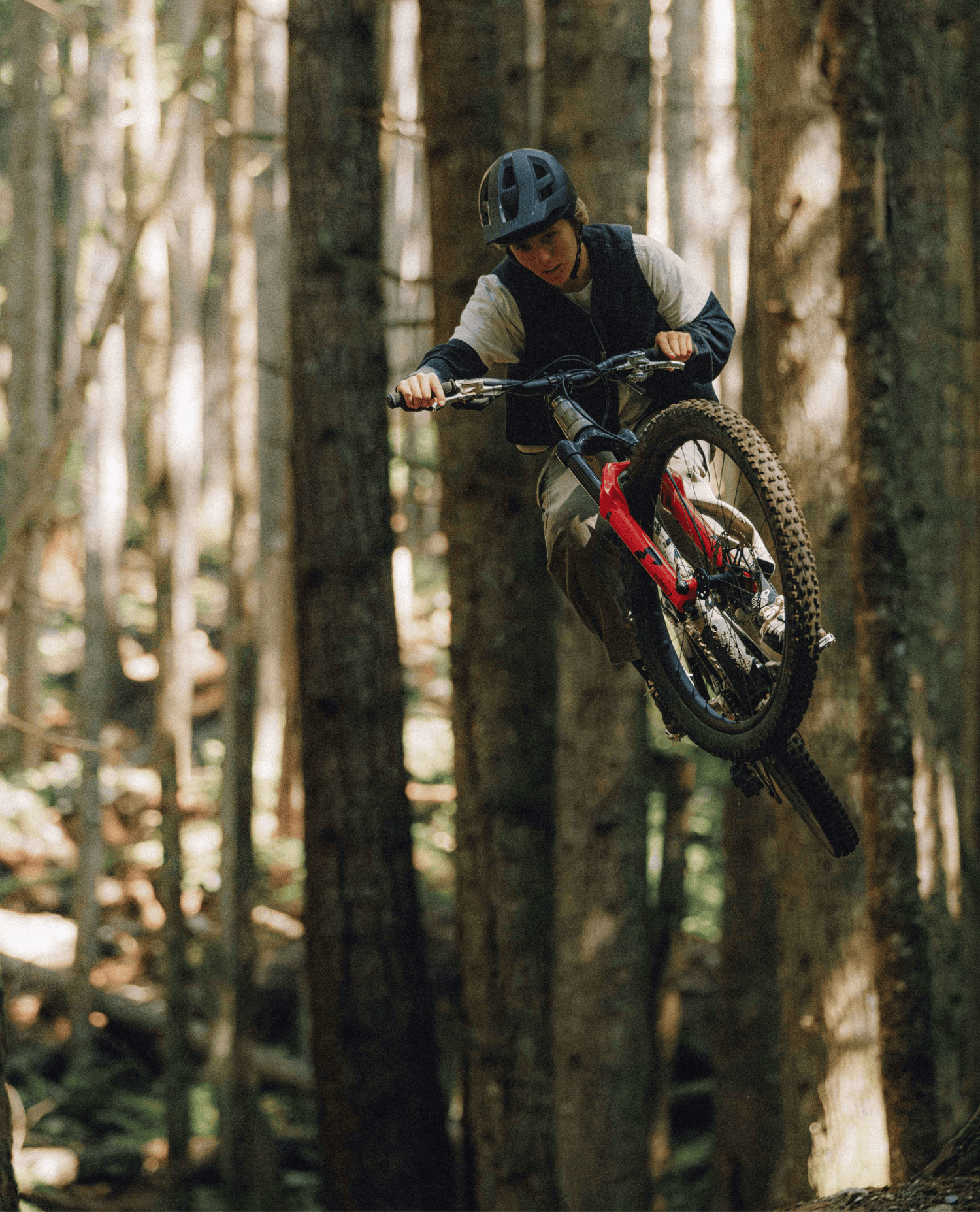 A mountain biker wearing a black helmet and riding gear jumps a red full-suspension mountain bike mid-air through a dense forest of tall tree trunks.