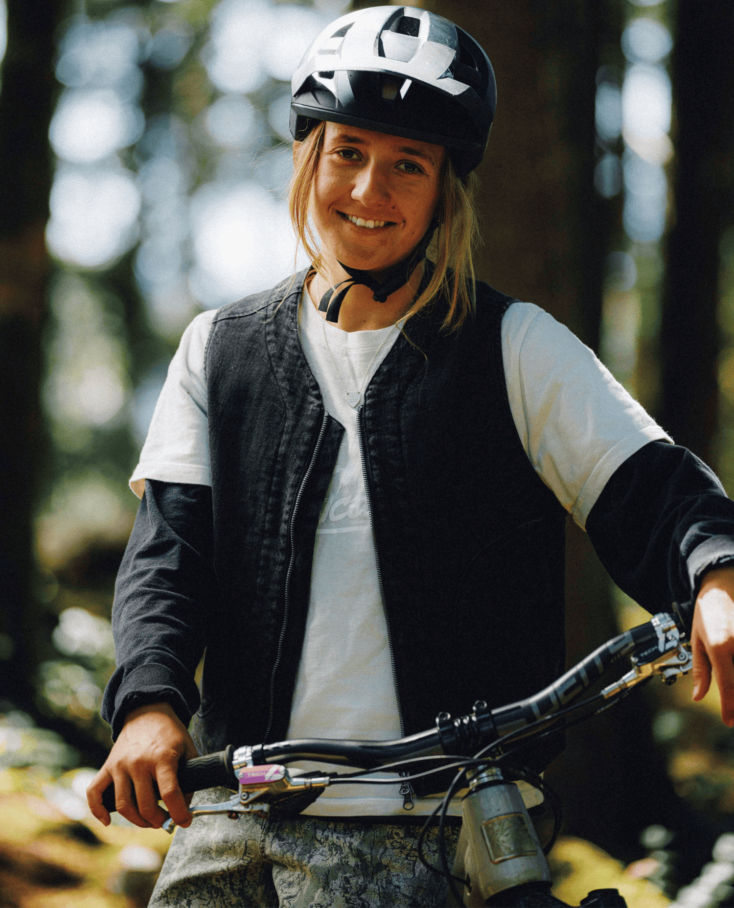 A smiling woman wearing a black and white mountain bike helmet, a black vest over a white long-sleeve shirt, and camouflage pants, posing with a bike in a forest.