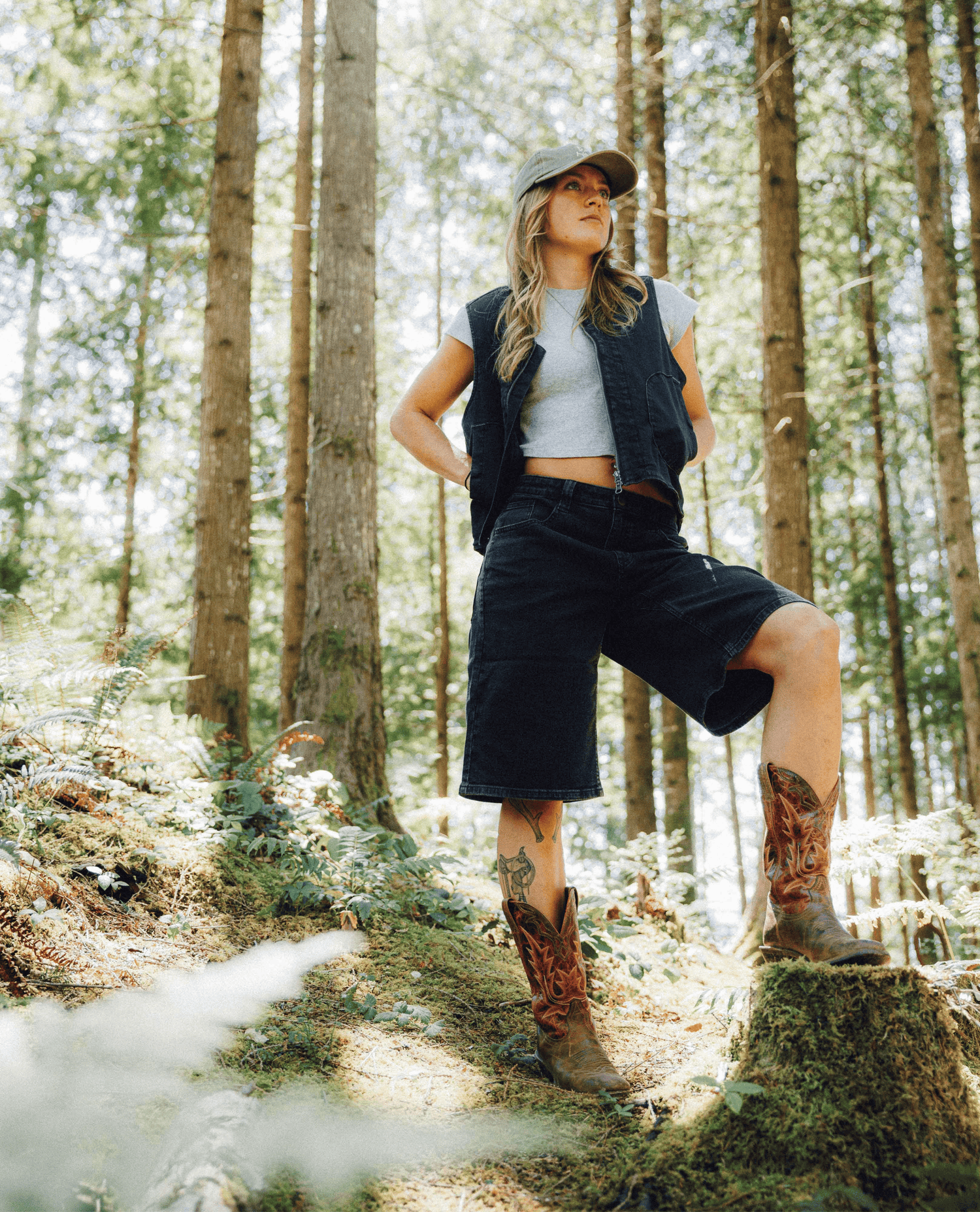 A woman stands on a mossy tree stump in a sunlit forest wearing a dark denim vest, matching shorts, a white cropped T-shirt, a baseball cap, and brown cowboy boots.