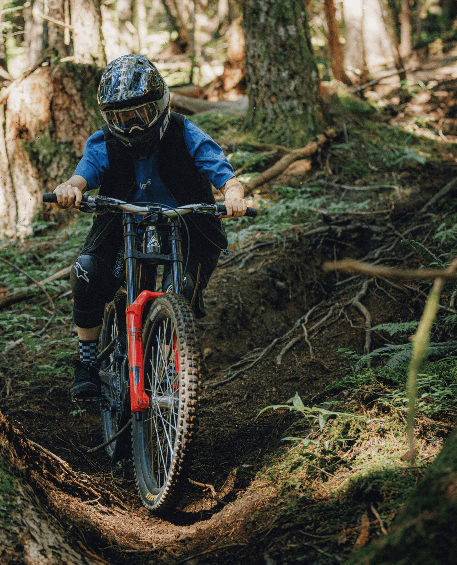 A mountain biker wearing a black helmet, blue shirt, and black pants rides a full-suspension bike with a bright red front fork through a forest trail.