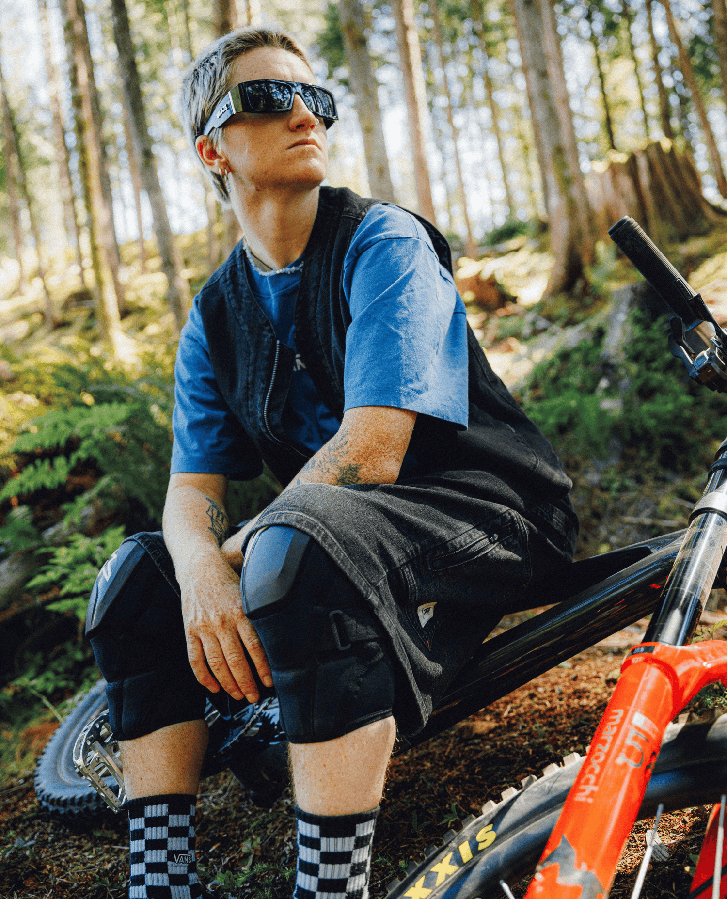 A person sitting on a mountain bike in a forest, wearing black sunglasses, a blue t-shirt, a black vest, and black shorts with knee pads. The person is also wearing black-and-white checkered socks, and an orange mountain bike frame is visible in the foreground.