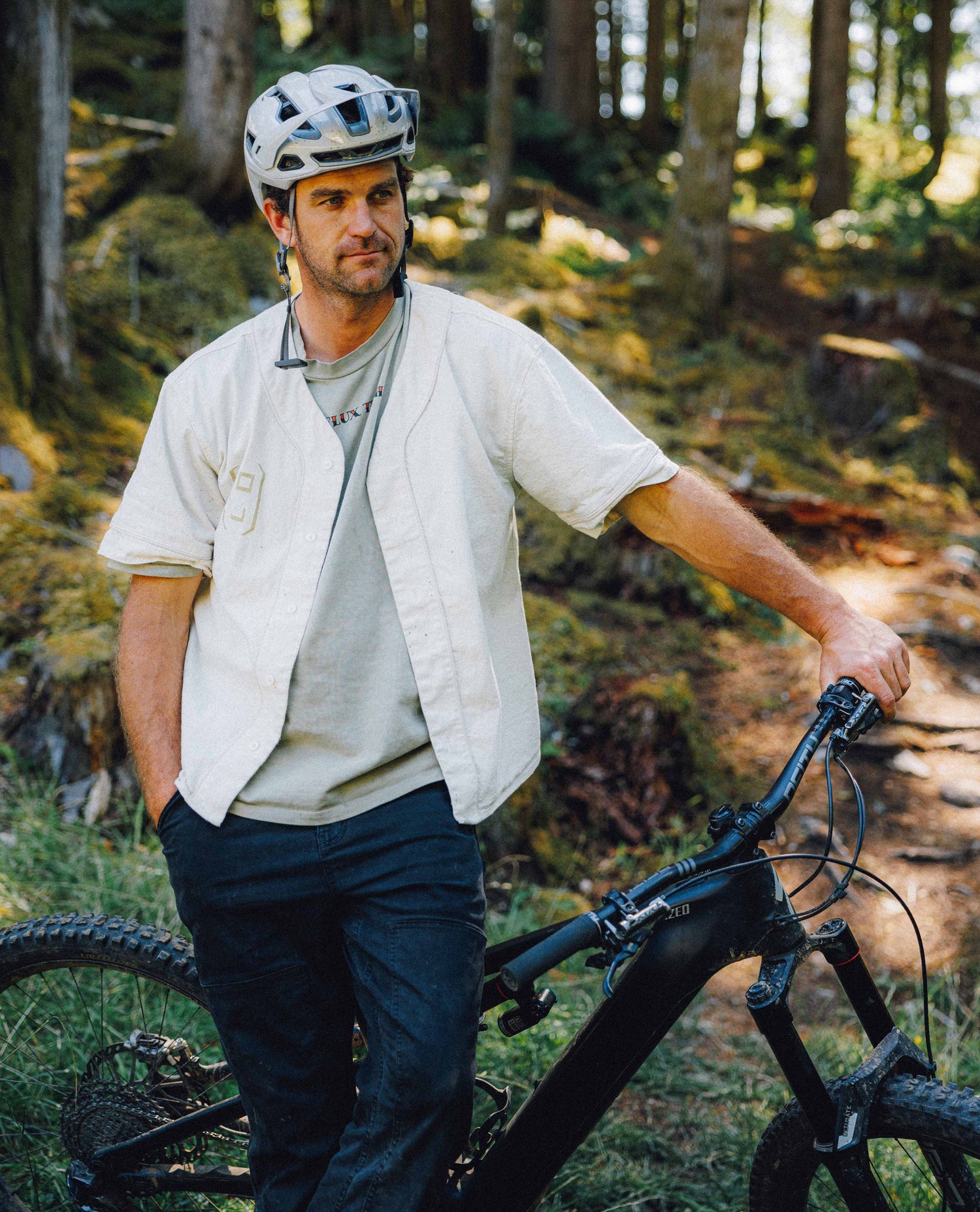 A man wearing a white helmet and a light beige short-sleeve button-up shirt stands next to a black mountain bike in a forest setting.