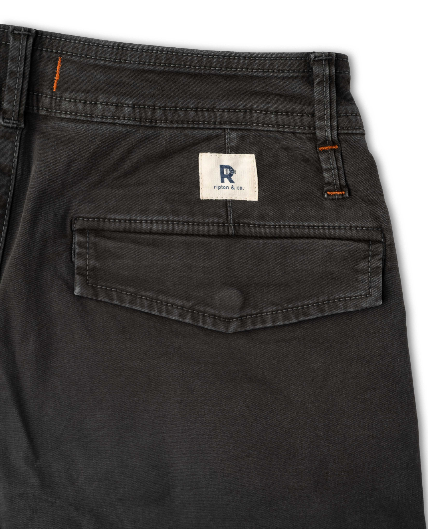 Close-up of the back waistband and pocket area of dark charcoal gray denim jeans, showing a white brand patch with a blue 'R' logo and orange contrast stitching.