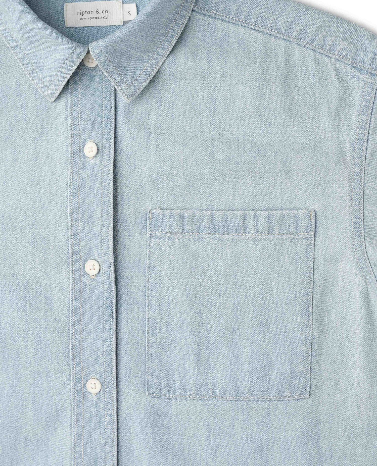 A close-up of a light blue chambray button-down shirt with a collar, white buttons, and a chest pocket. The brand label 'Ripton & co.