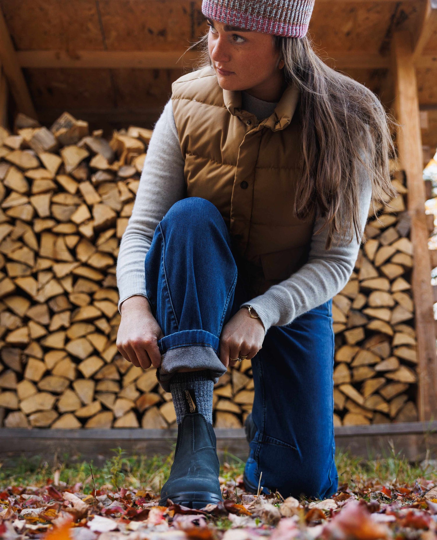 Person sitting in front of stacked firewood wearing a brown vest, blue jeans, and a pink knit hat.
