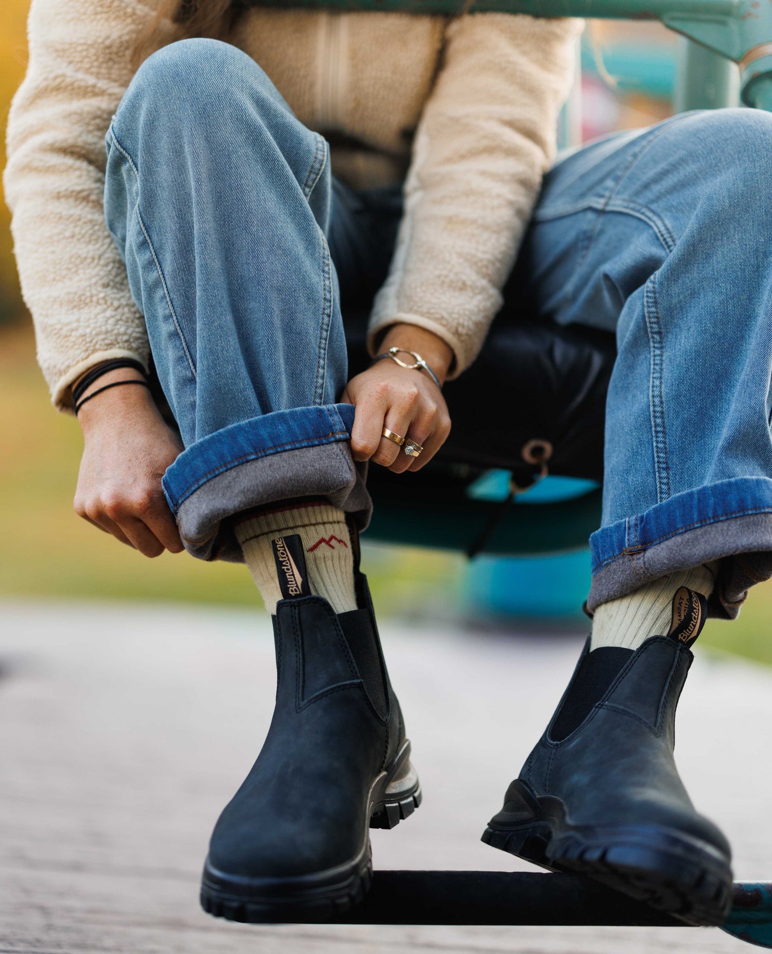 Person wearing blue Chelsea boots, rolled-up jeans, and patterned socks with a blurred outdoor background.