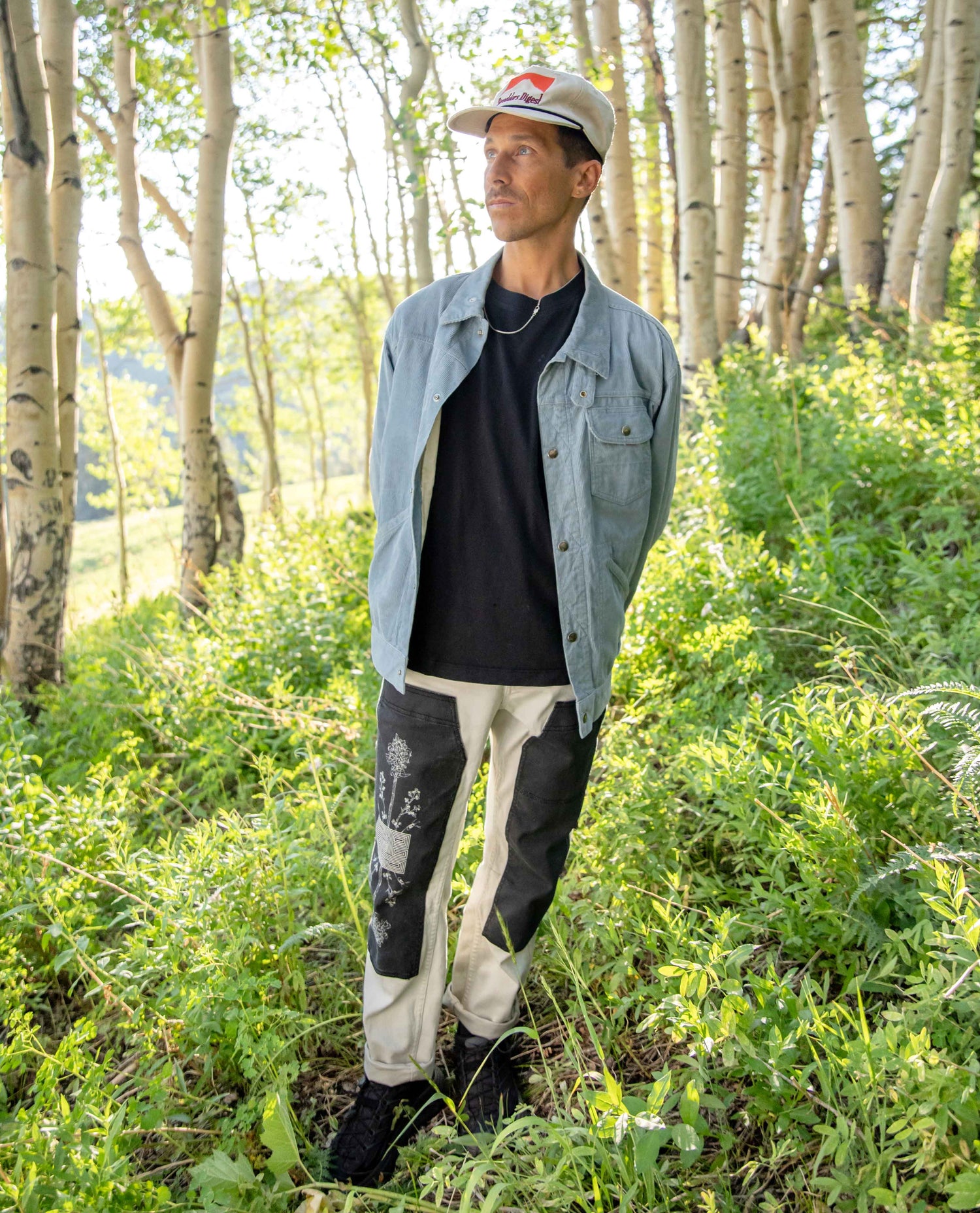 A man standing in a sunlit forest clearing wearing a light blue denim overshirt, black t-shirt, distressed off-white and black pants, and a beige baseball cap.
