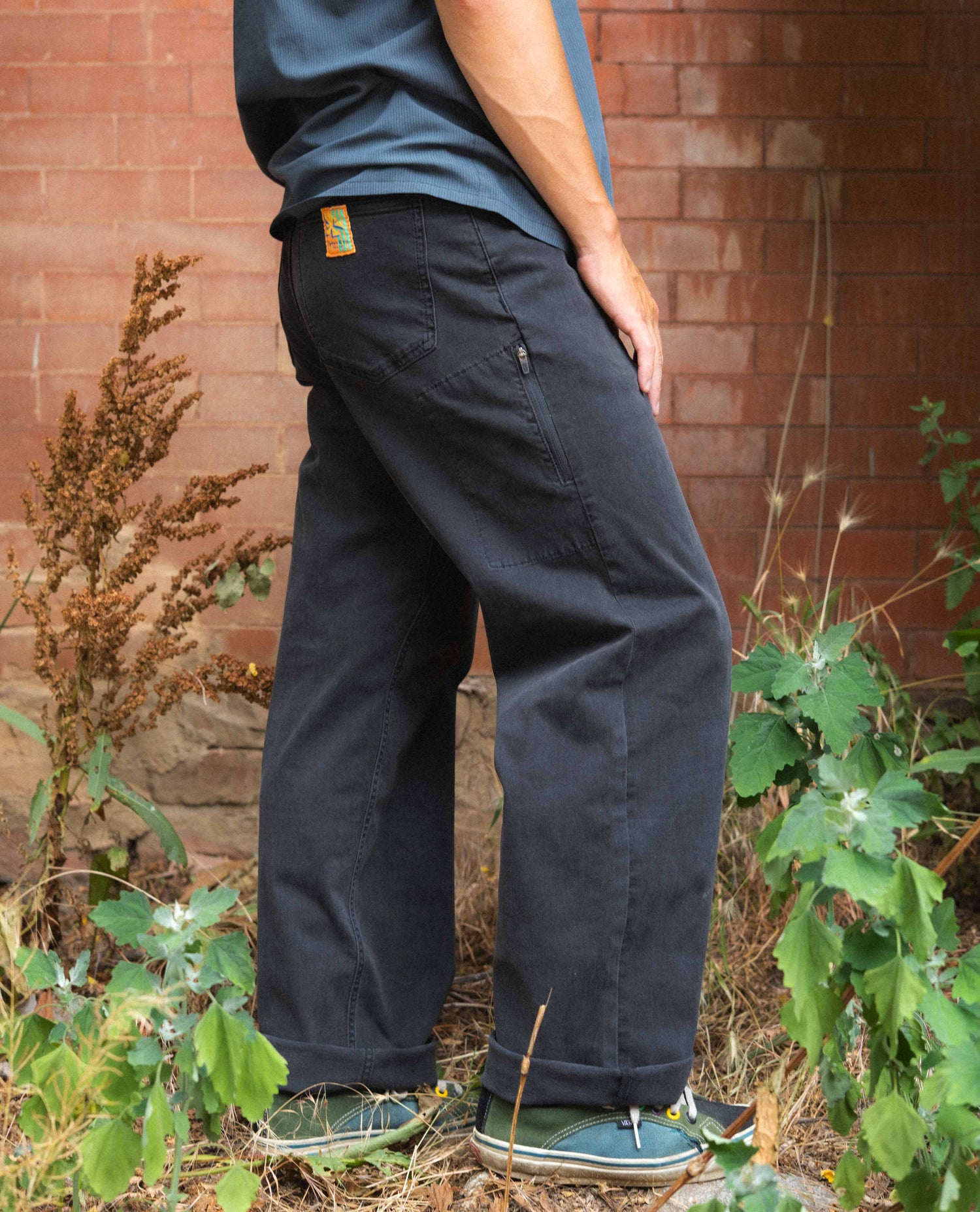 Person wearing dark pants and a blue shirt standing in front of a brick wall with plants around.