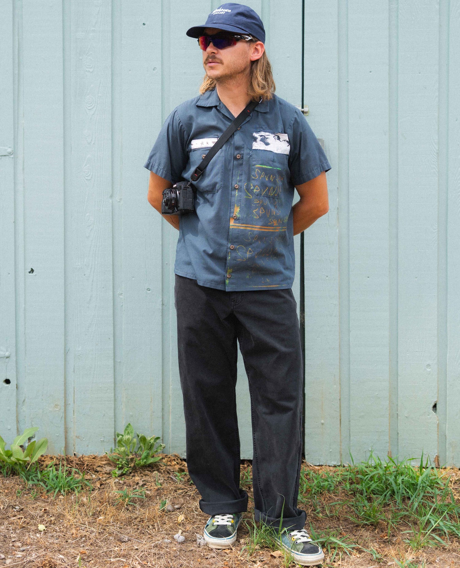 Person wearing a blue denim shirt and black pants standing against a light blue wall.