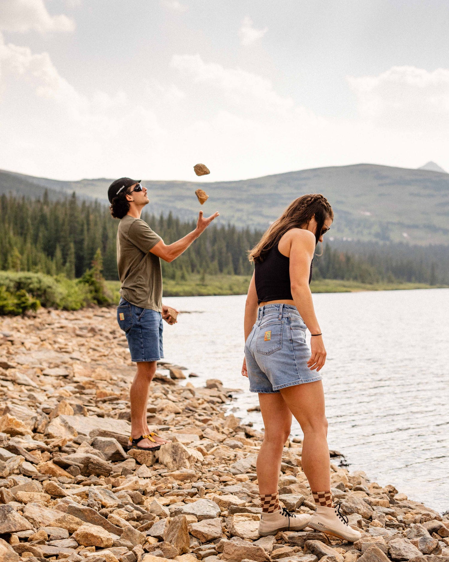 Woman standing near a mountain lake in Ripton jorts with a man in the background juggling some rocks. 