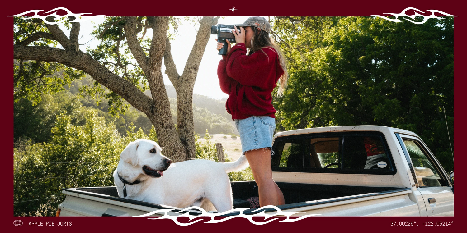 A woman in a red sweatshirt and denim shorts stands in the bed of a white pickup truck taking a photo with a camera, while a white dog sits in the truck bed. The scene is outdoors with trees and hills in the background, framed by a dark red border with decorative flame-like corner flourishes and small text at the bottom.