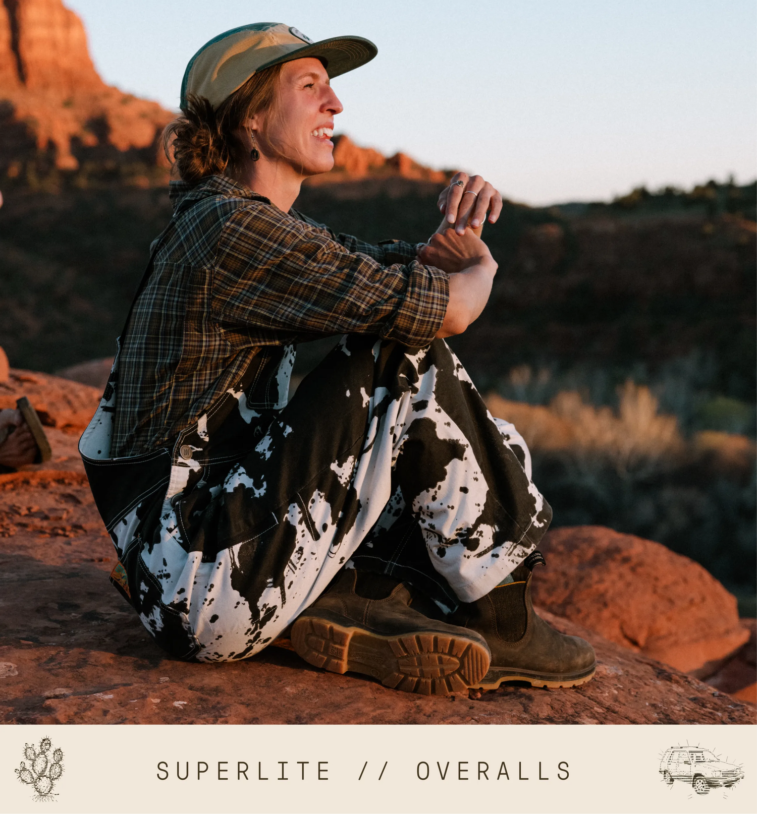 Person sitting on a rock with scenic desert background, wearing plaid shirt and patterned overalls.