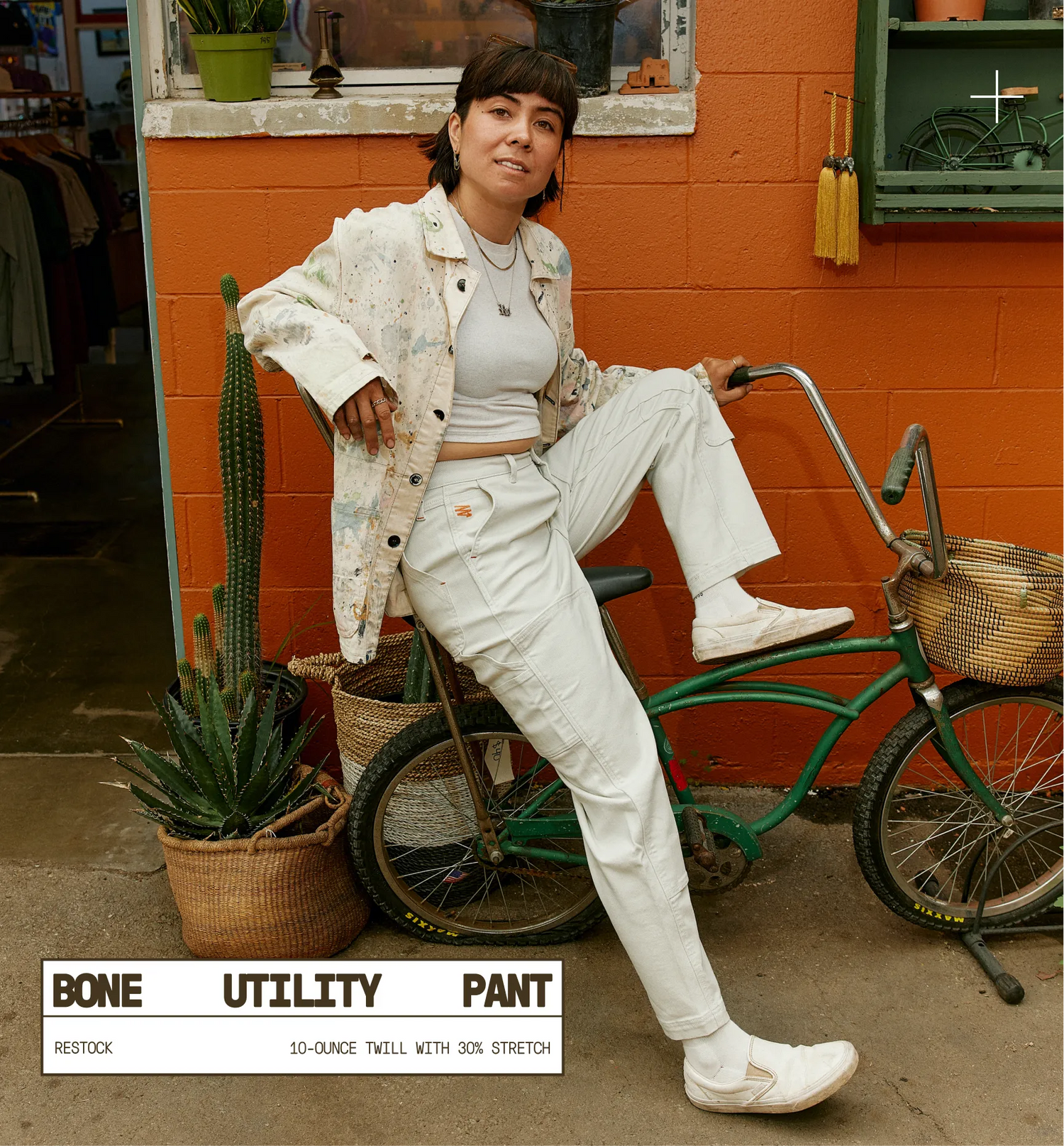 Woman in a white outfit sitting on a bicycle against an orange wall with plants.